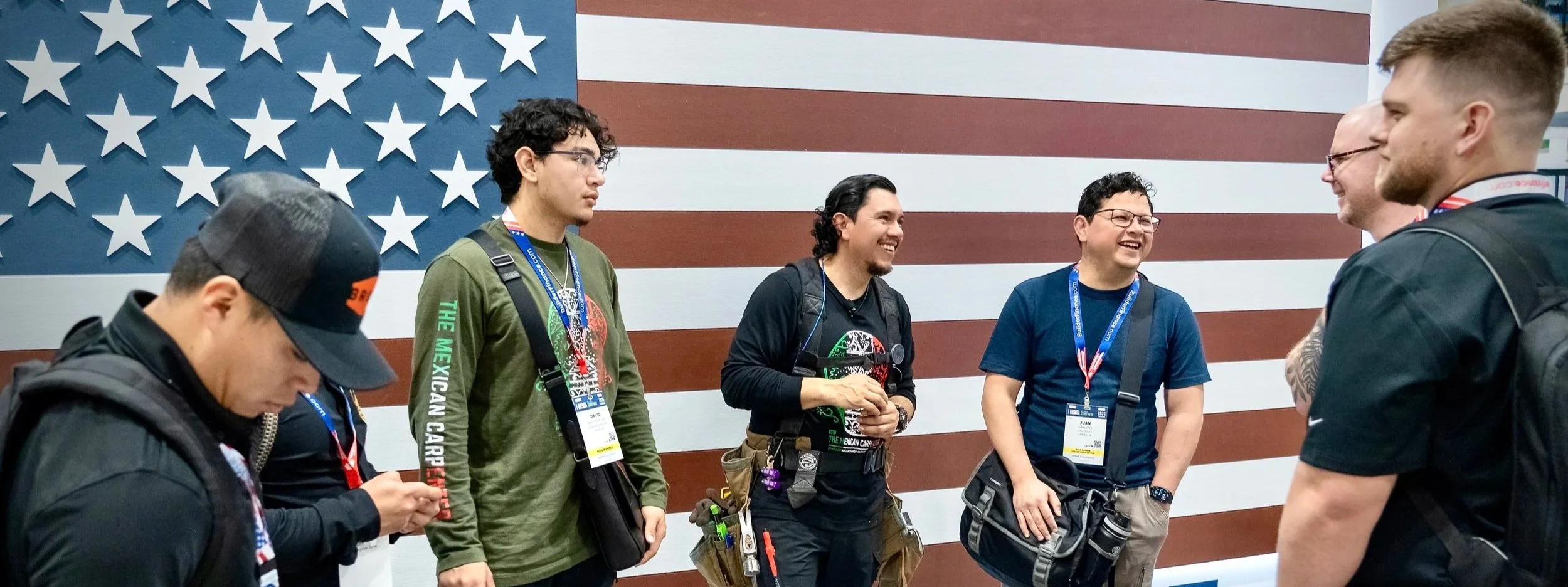 A group of five men standing and chatting in front of an American flag wall at an event, with some wearing backpacks and lanyards. Excited about  Latino builders show taking place September 24th 2026 at the Baltimore convention center in Maryland