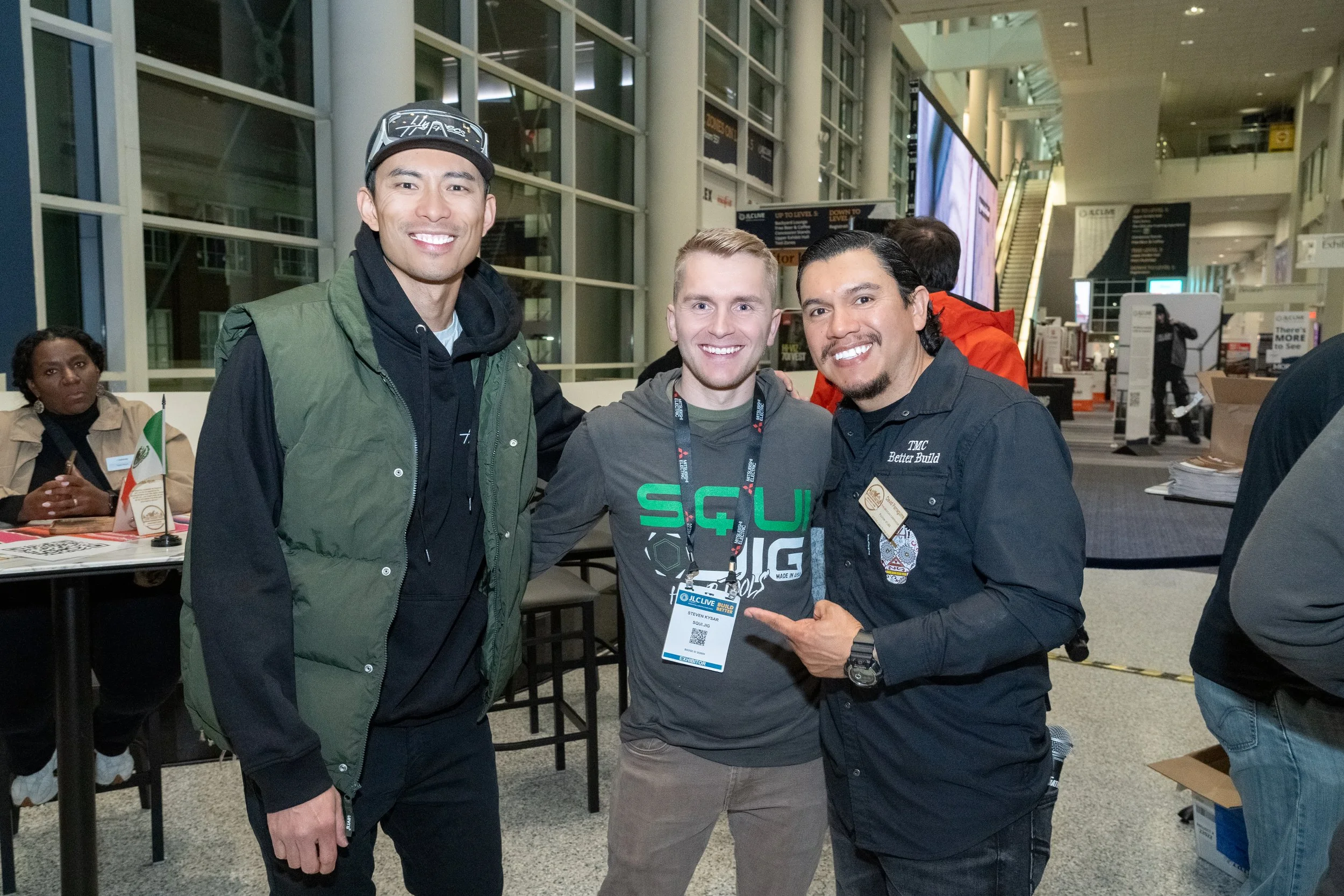 Three men standing together at an indoor event, smiling at the camera. The man on the left is wearing a baseball cap, black hoodie, and green vest. The man in the middle is wearing a gray T-shirt with green and black lettering and has a lanyard with a badge. The man on the right is wearing a black shirt with a badge, pointing at the man in the middle.
