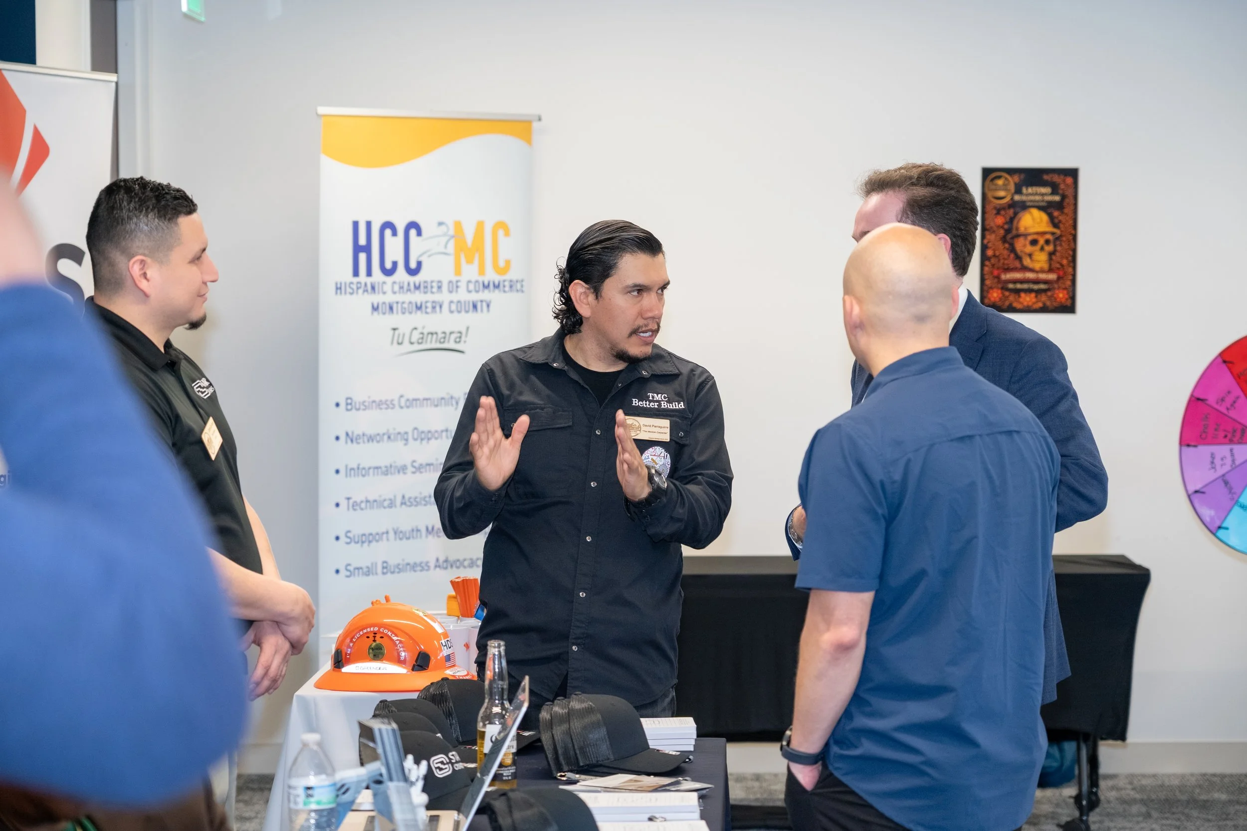 A man in a black shirt explaining something to a group of three men at a professional event. There is a table with a hard hat, hats, and bottled drinks. A banner for the Hispanic Chamber of Commerce in Montgomery County is visible in the background, along with some artwork on the wall.