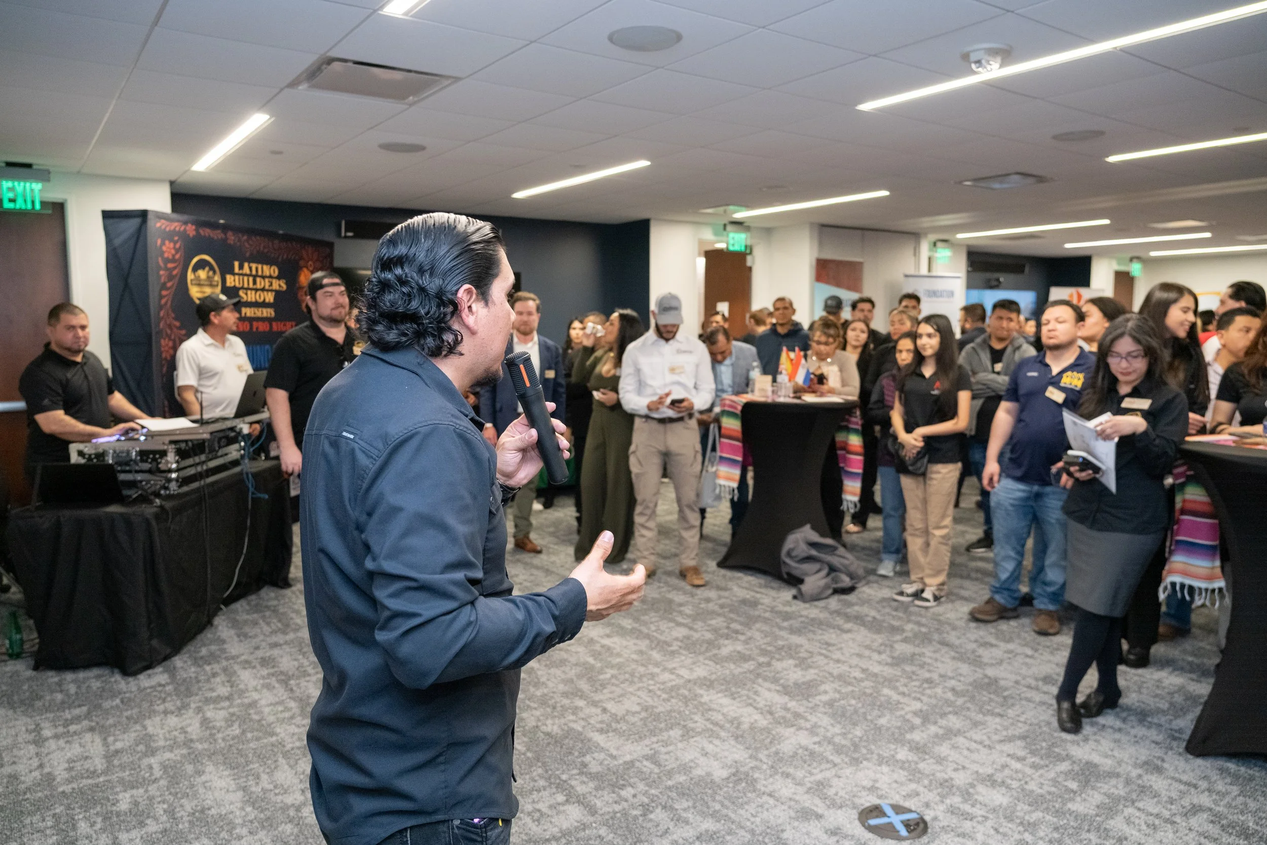 A man with dark, wavy hair is speaking into a microphone in front of a large group of people at an indoor event, with a banner in the background that reads "Latino Builders Show." The audience is standing around high-top tables, some taking notes or pictures.