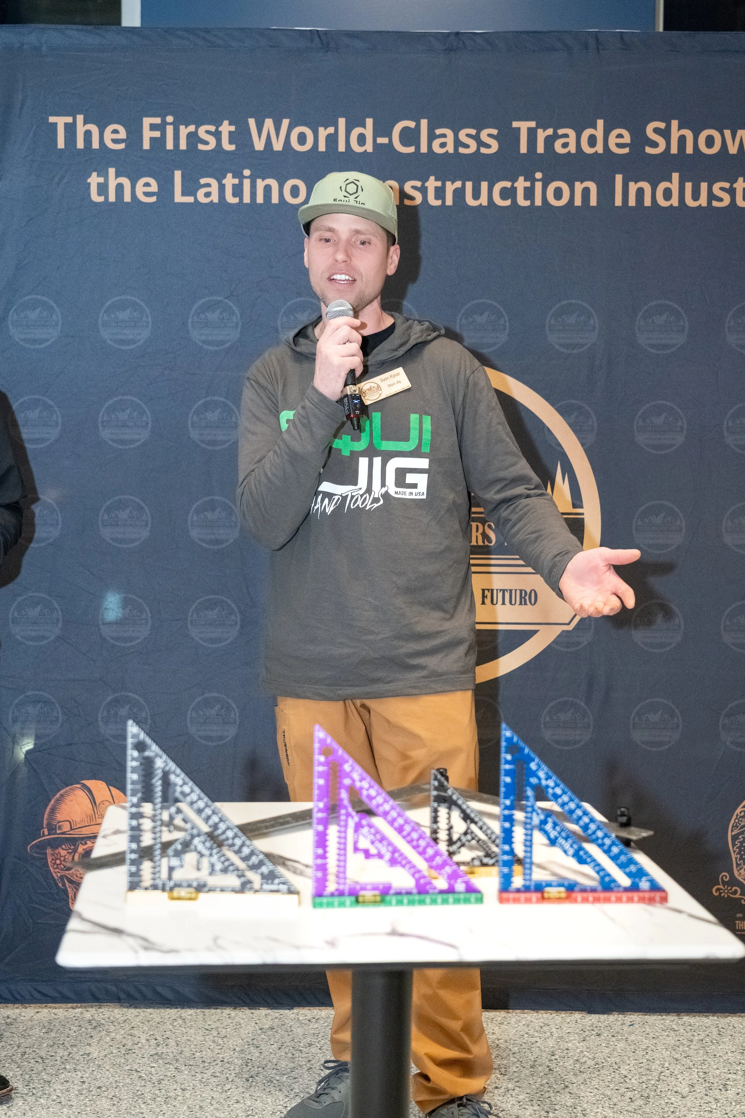 A man is talking into a microphone at a trade show in front of a dark backdrop with text about a trade show for the Latino construction industry. There are colorful measuring tools or triangles on a table in front of him.