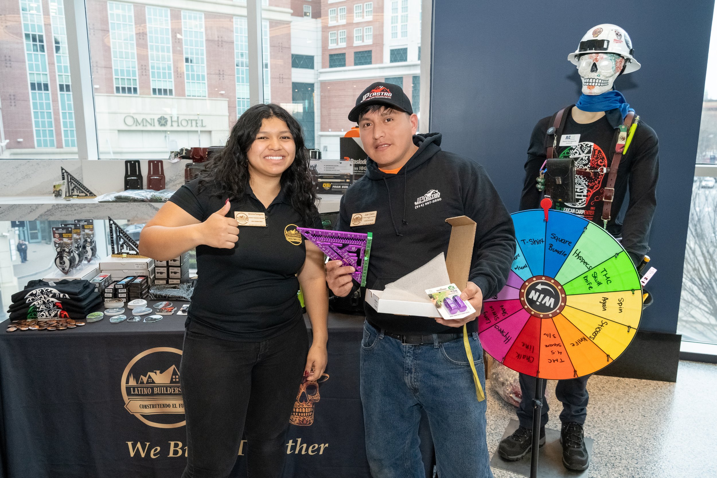 Two people standing inside an event space, one woman with dark curly hair giving a thumbs-up and smiling, and a man holding a box with purple items, next to a colorful spin wheel. Behind them is a table with merchandise and a skeleton figure on display.