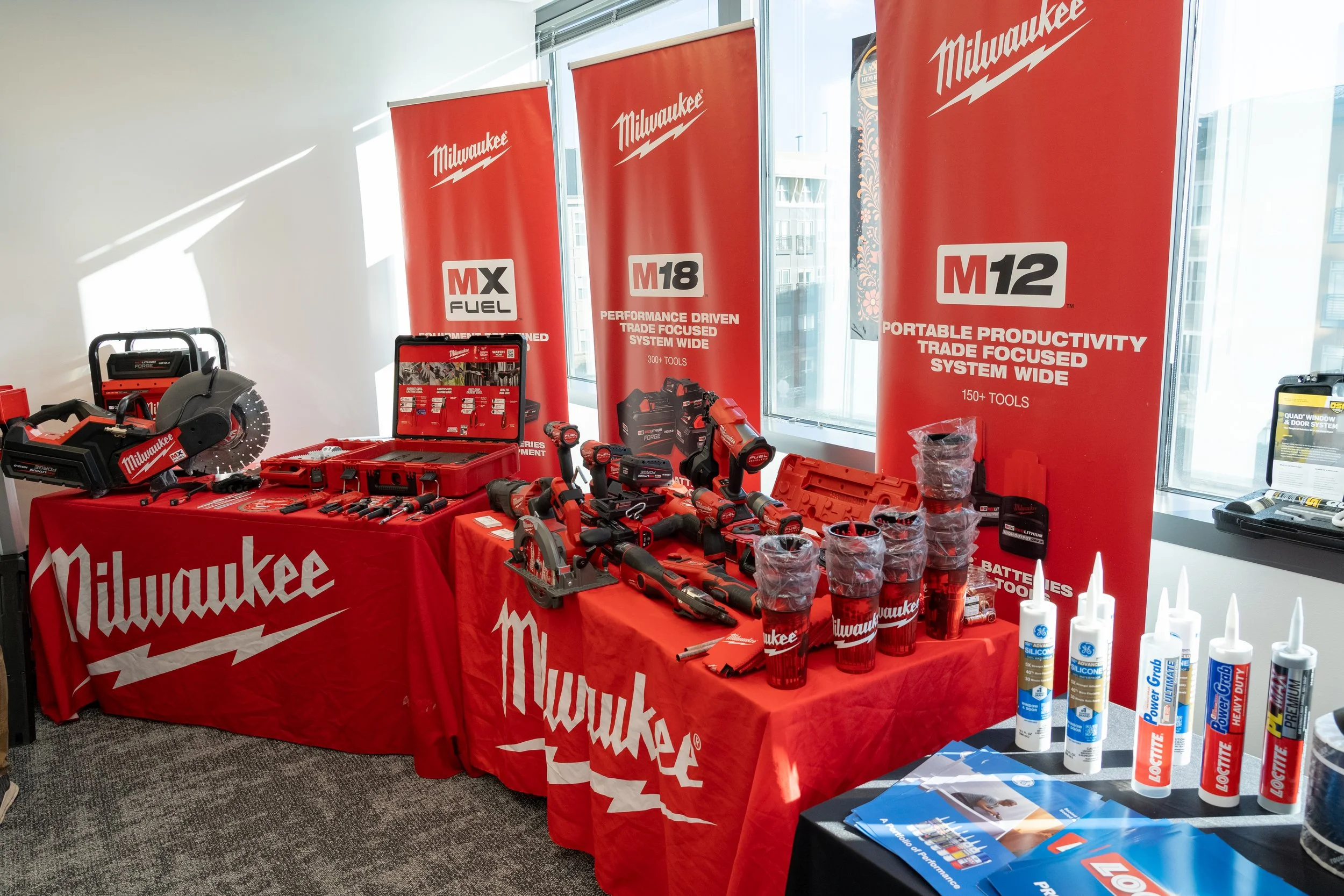 Display of Milwaukee power tools including saws and drills, with red banners and tables, brand logos, and storage bottles in the background.