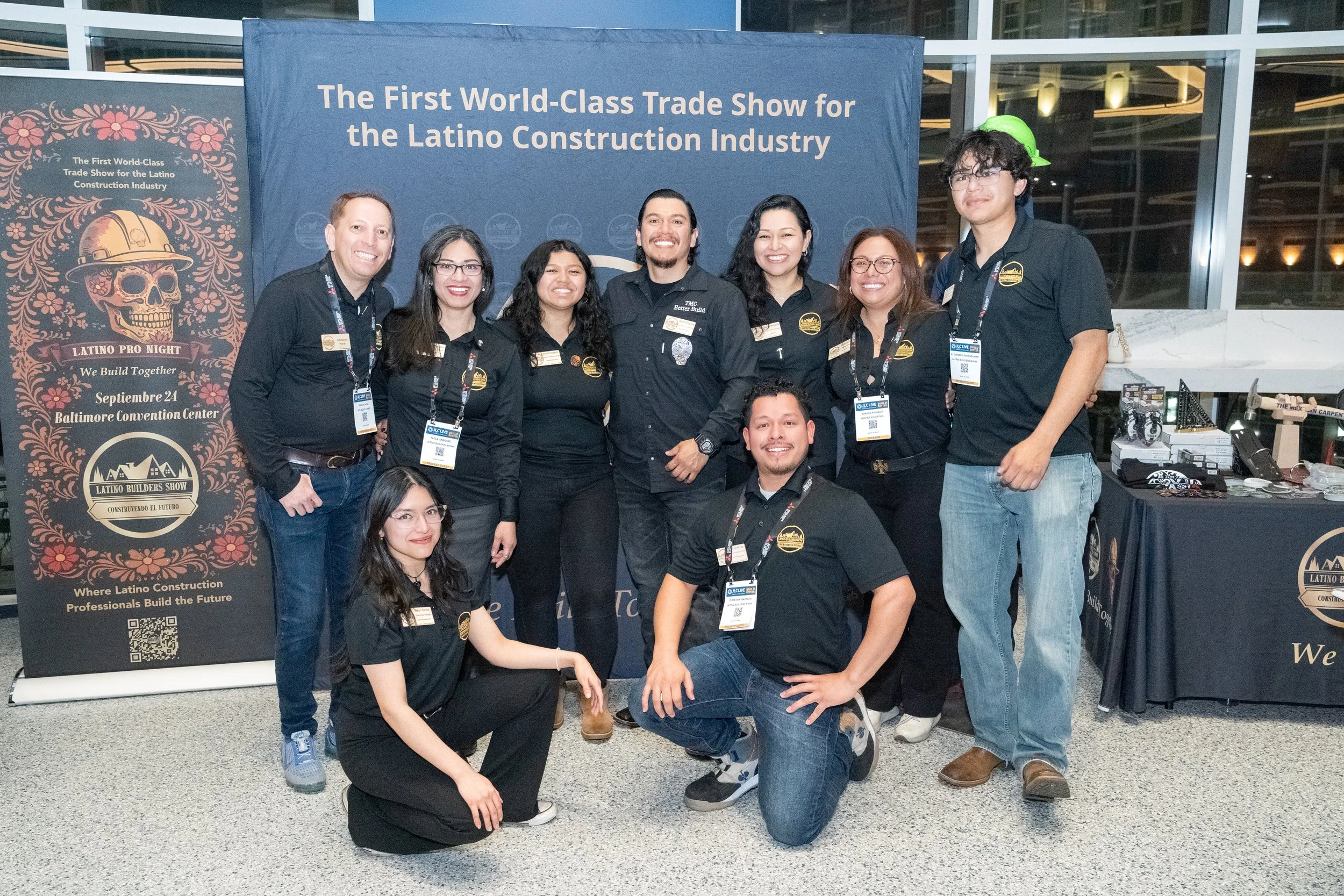 Group of nine people at a trade show for the Latino construction industry, standing in front of a blue banner that reads 'The First World-Class Trade Show for the Latino Construction Industry.' The group is smiling and wearing black shirts with a logo, and some are wearing lanyards with badges.