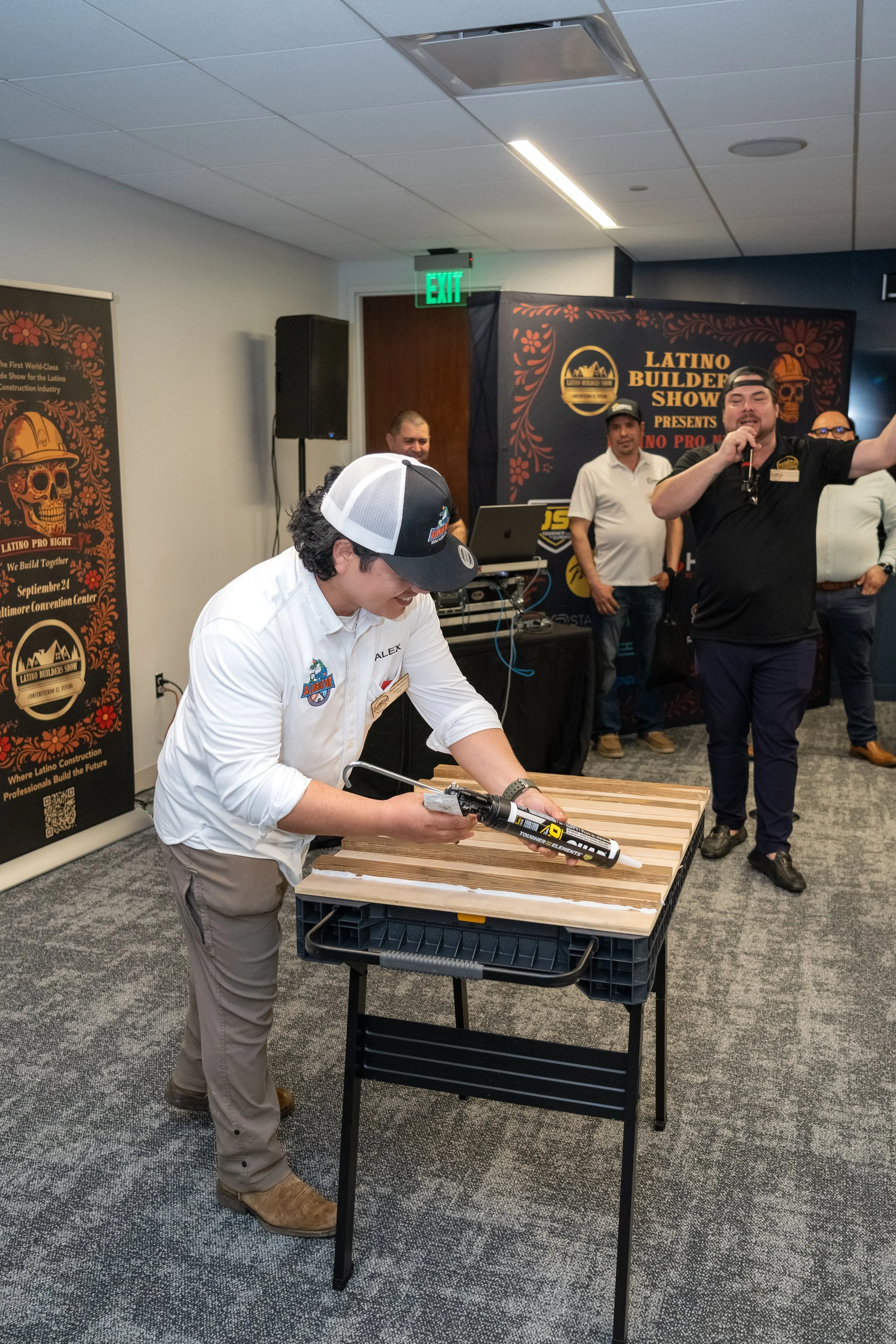 A man in a white shirt and cap is applying construction adhesive to wooden planks on a table at a building industry event, with a group of people observing and a banner in the background reading "Latino Builder Show".