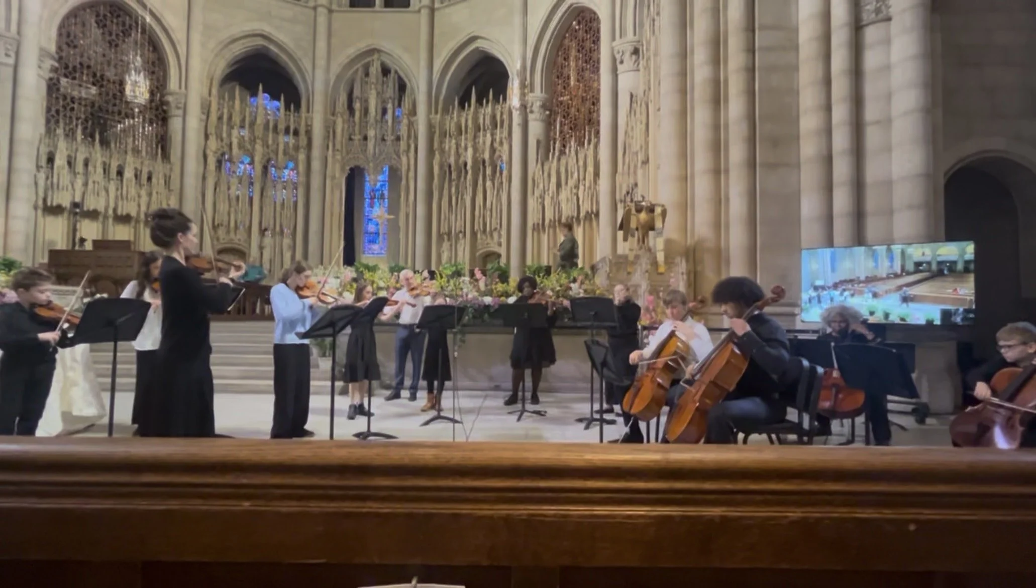 A group of children and adults playing violins and cellos during a performance inside a grand cathedral with high arched ceilings, stained glass windows, and ornate woodwork.