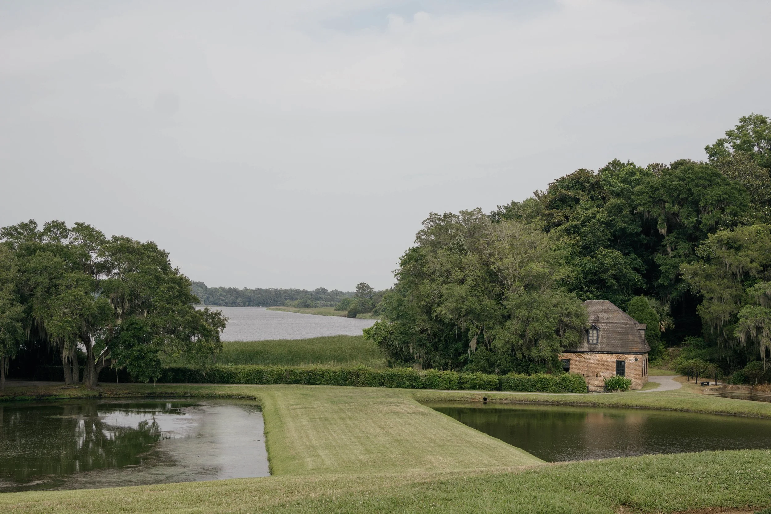 A serene landscape with a small brick house surrounded by greenery, two ponds with grass borders, trees, a waterway in the distance, and a cloudy sky.