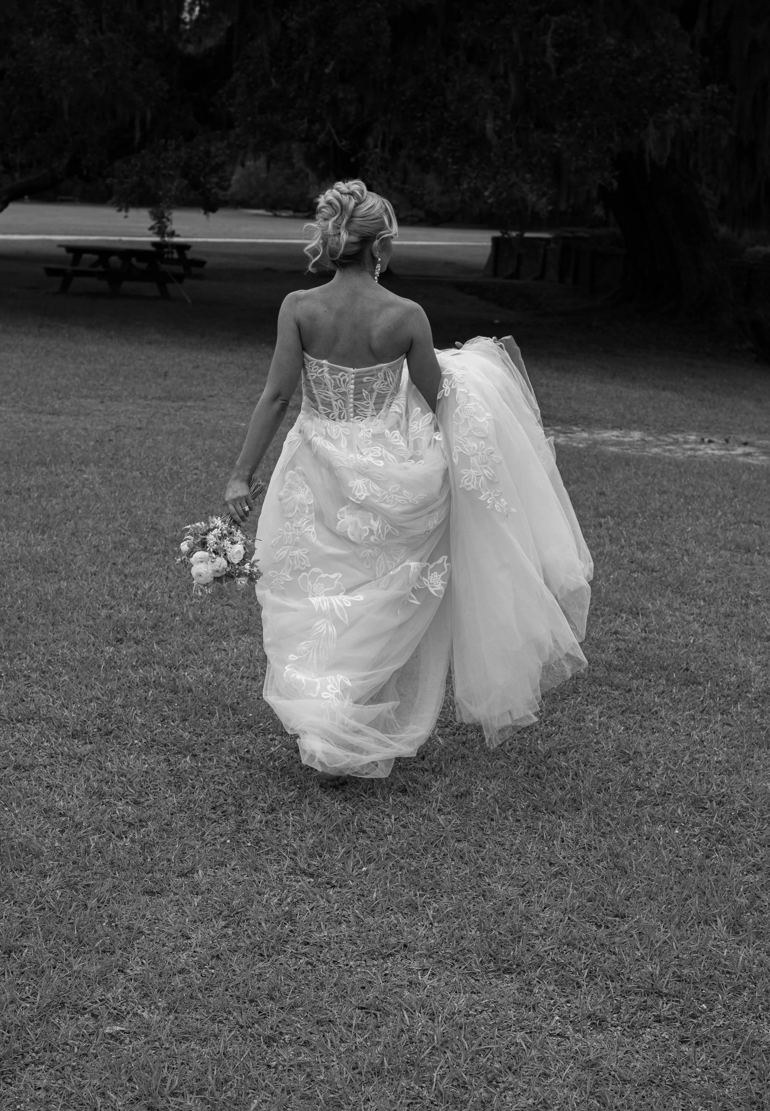 A bride in a strapless wedding gown holding a bouquet of flowers, walking on grass in a park with trees in the background.