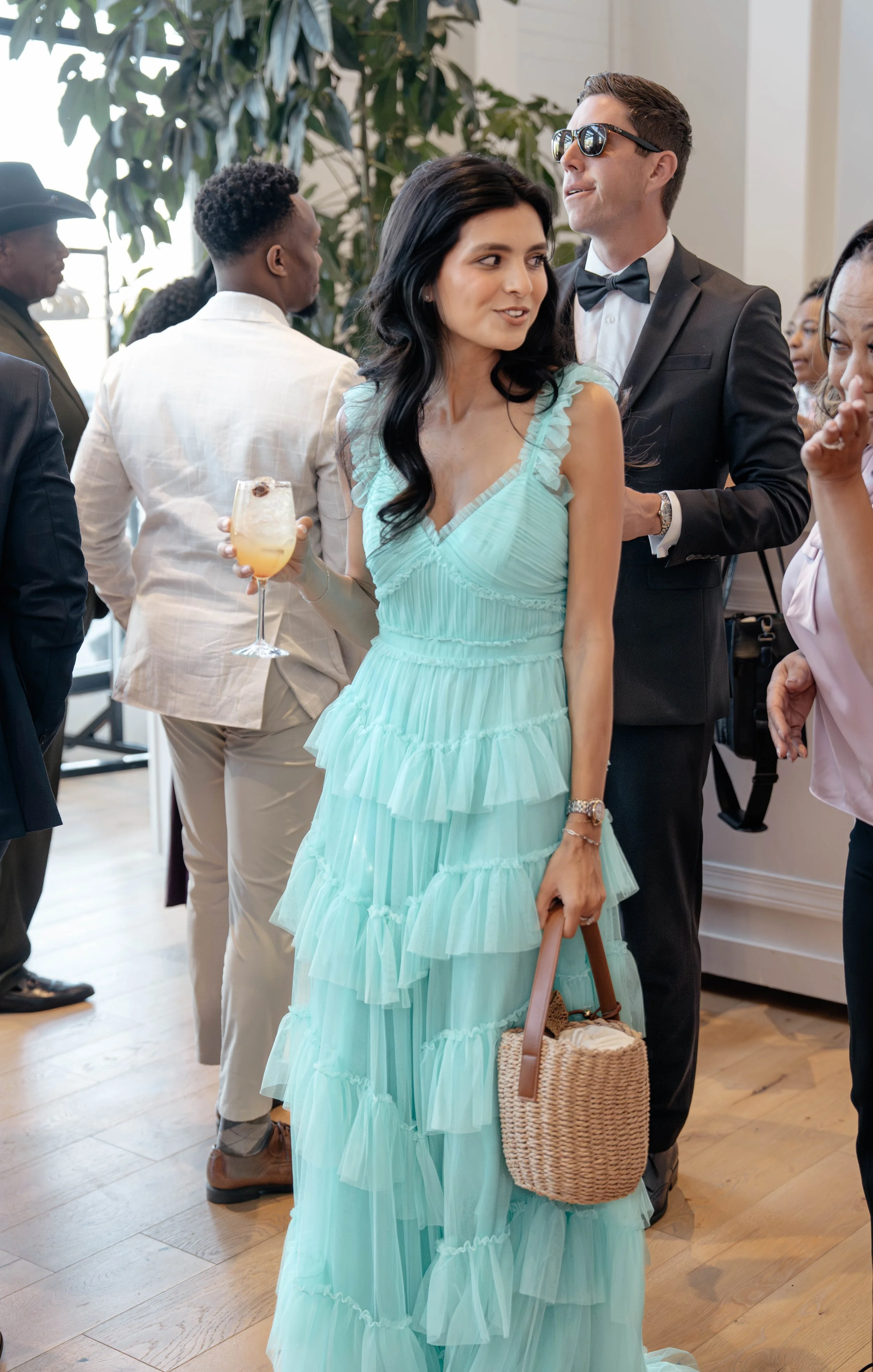 A woman in a light blue, ruffled, tiered dress holding a wine glass and a woven basket purse at a social event, with people in formal attire in the background.