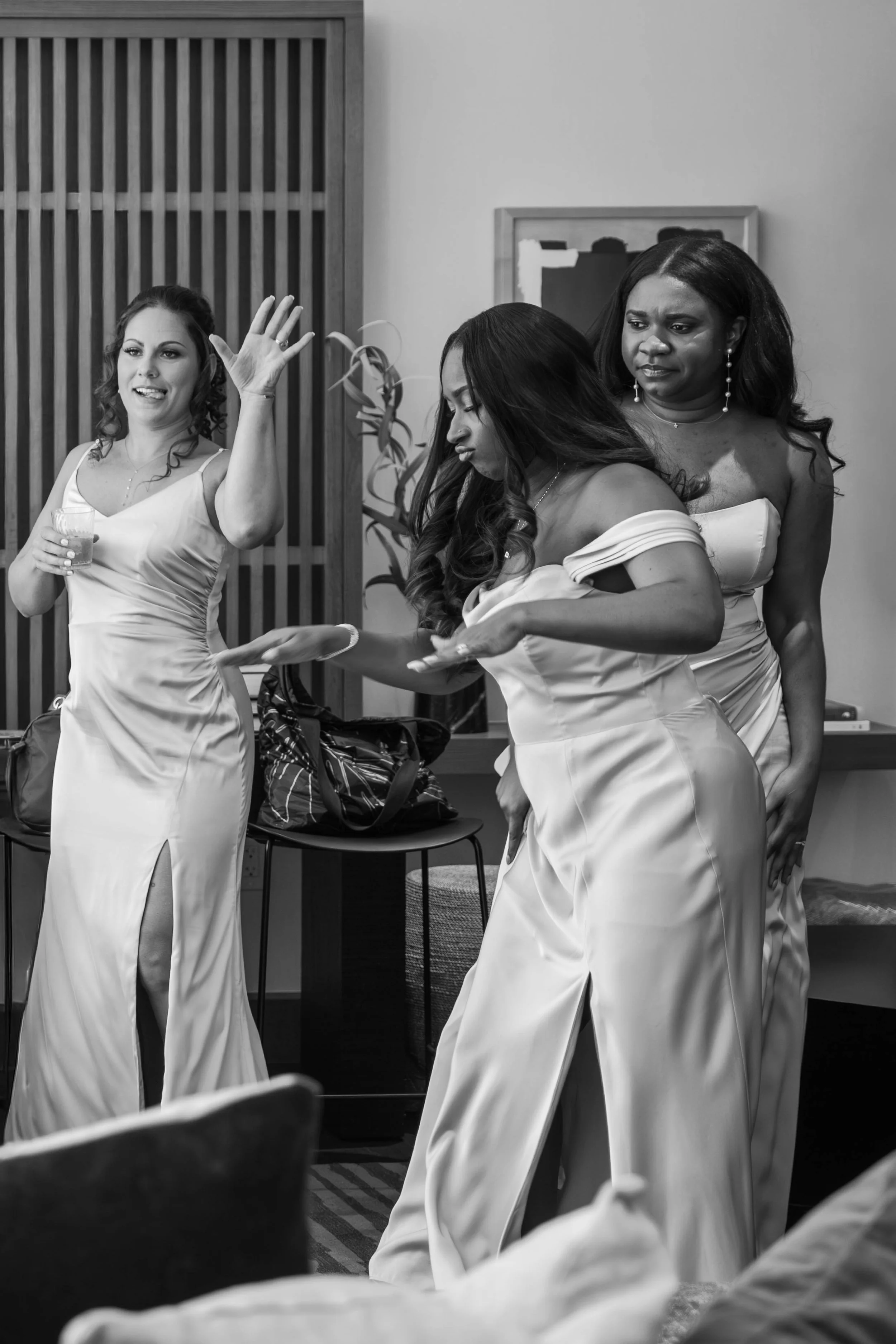 Three women in satin dresses at a social gathering, with one holding a drink and making a gesture, with indoor decor and artwork in the background.
