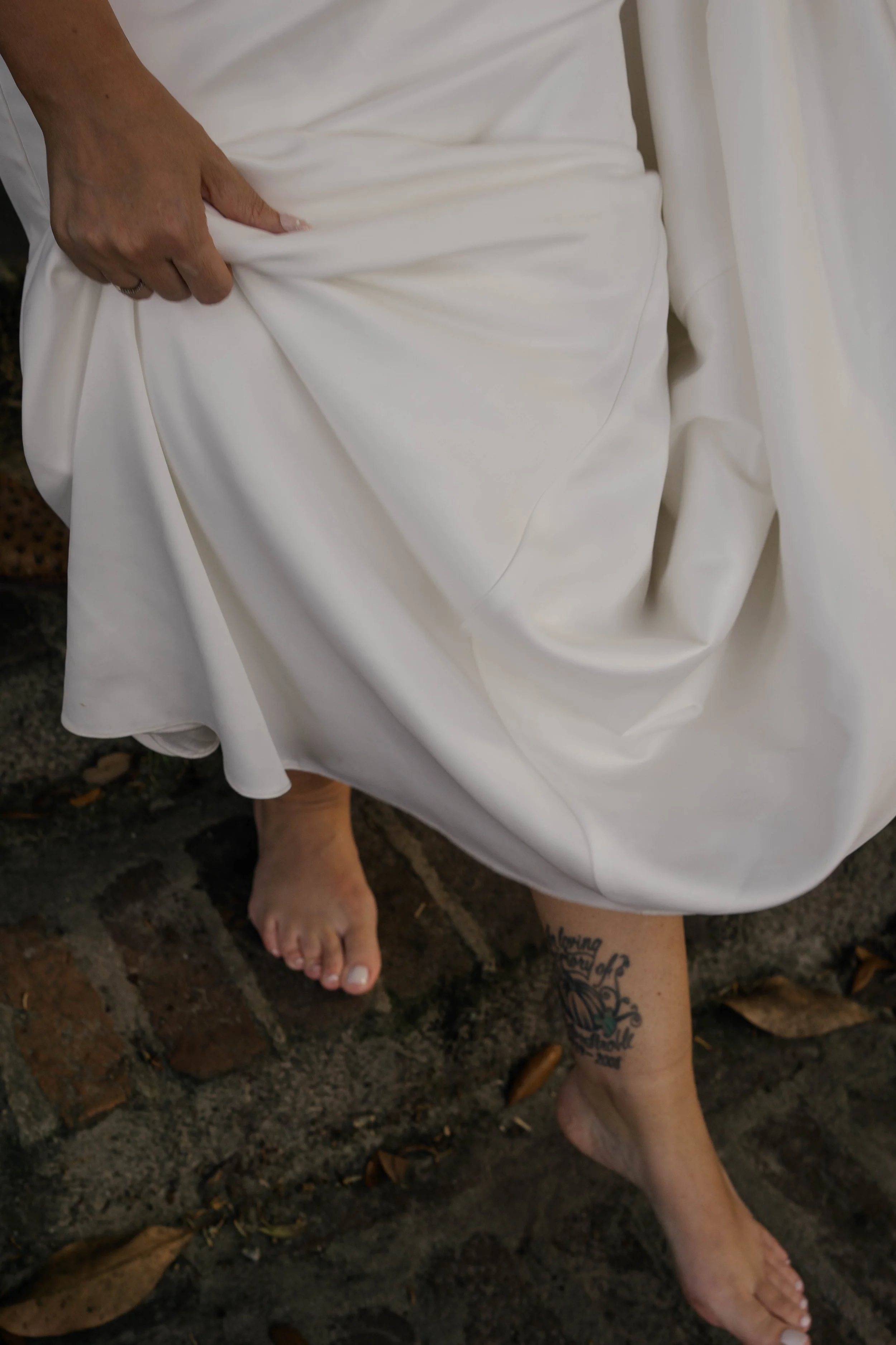 Person in a white dress lifting the fabric with one hand, standing barefoot on a brick and concrete surface. A tattoo is visible on their lower leg.