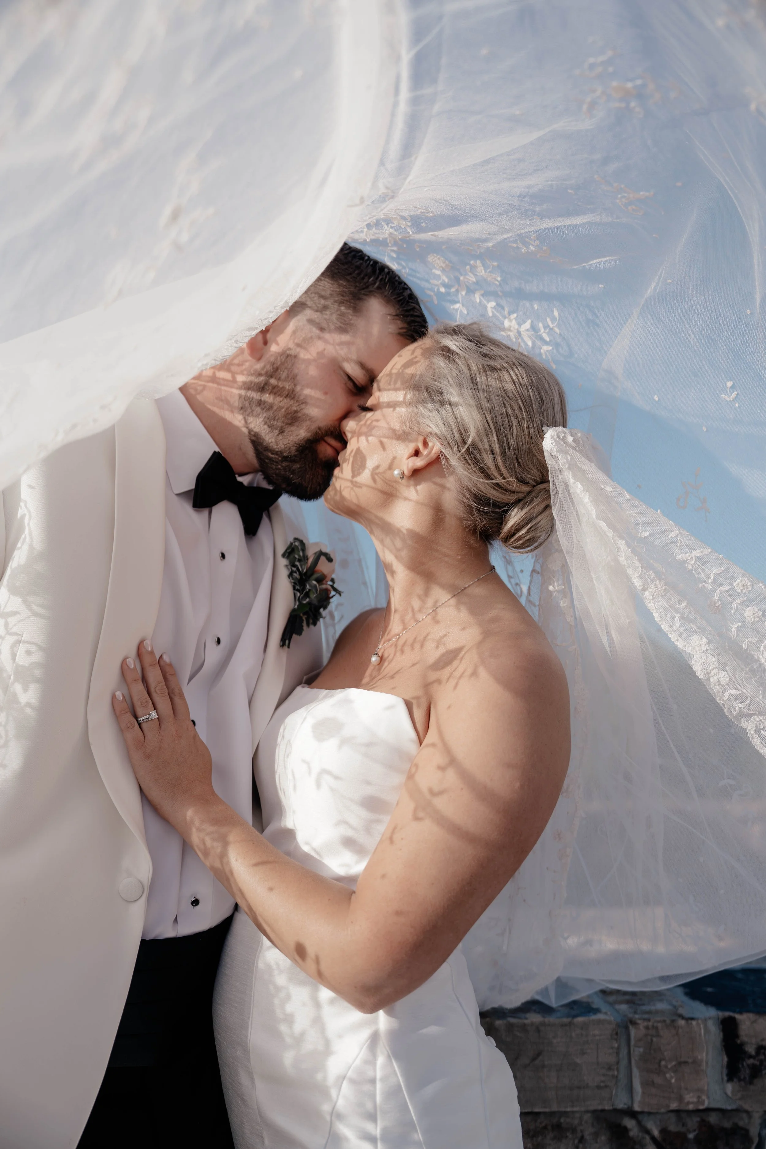 A bride and groom sharing a tender kiss under a sheer veil, with shadows of leaves on their faces.