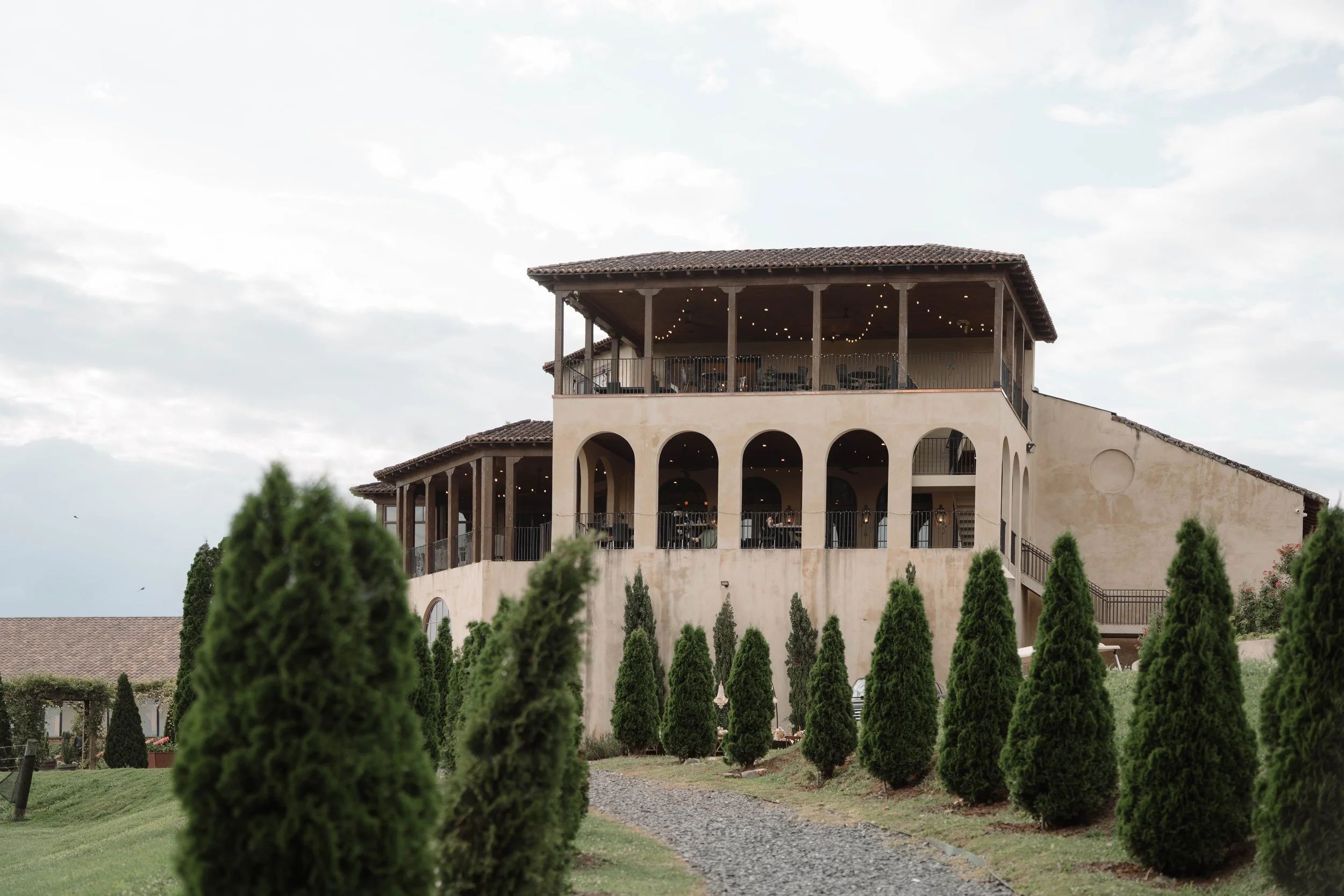 A multi-story building with arched openings, a balcony with string lights, and surrounding greenery and trees.