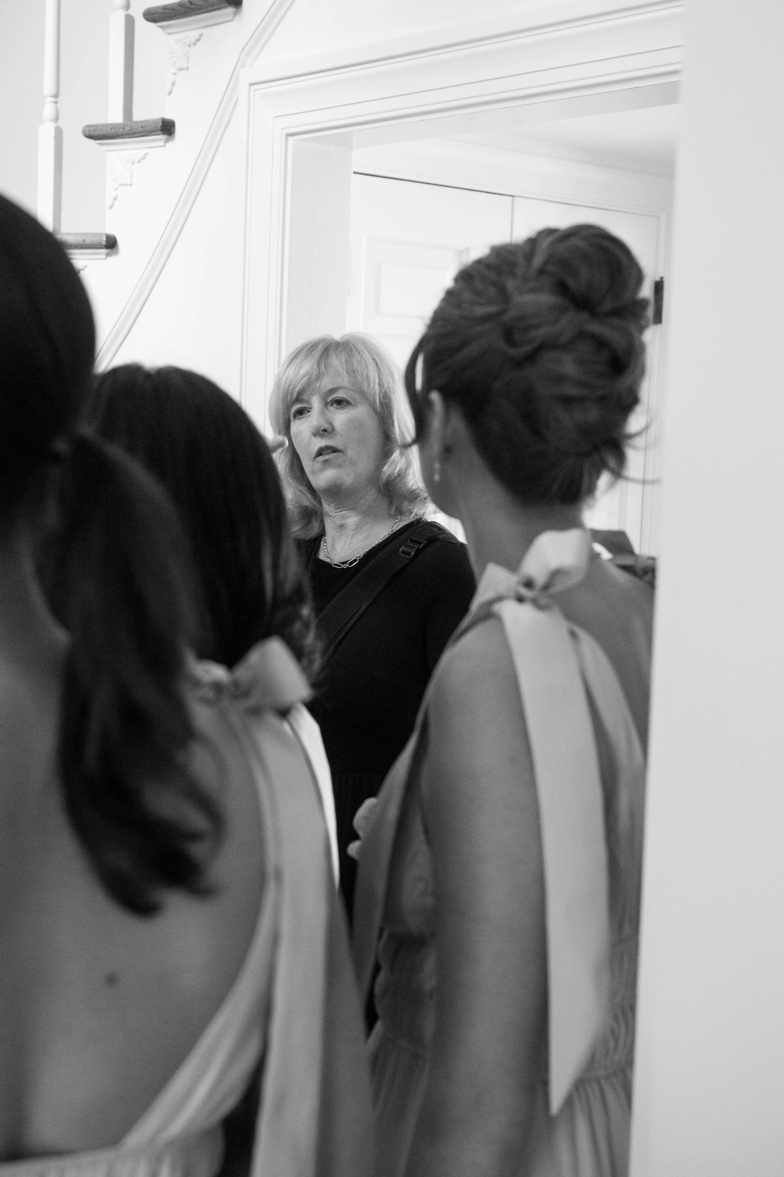 A woman with blonde hair and a dark top looking at a group of young women in formal dresses by a mirror in a room with white walls and crown molding.