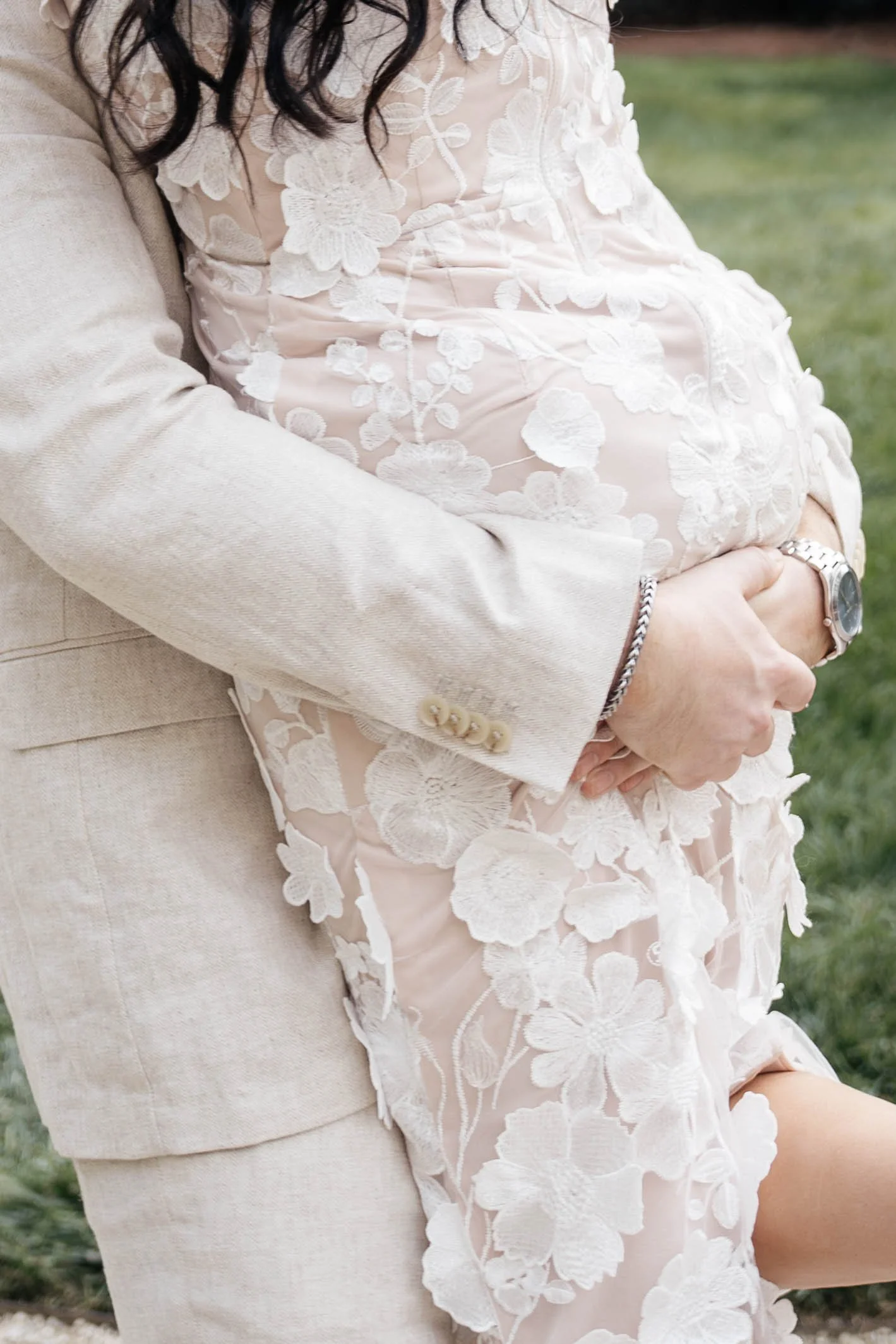 A pregnant woman wearing a white floral lace dress and a man wearing a beige suit jacket holding her belly outdoors.