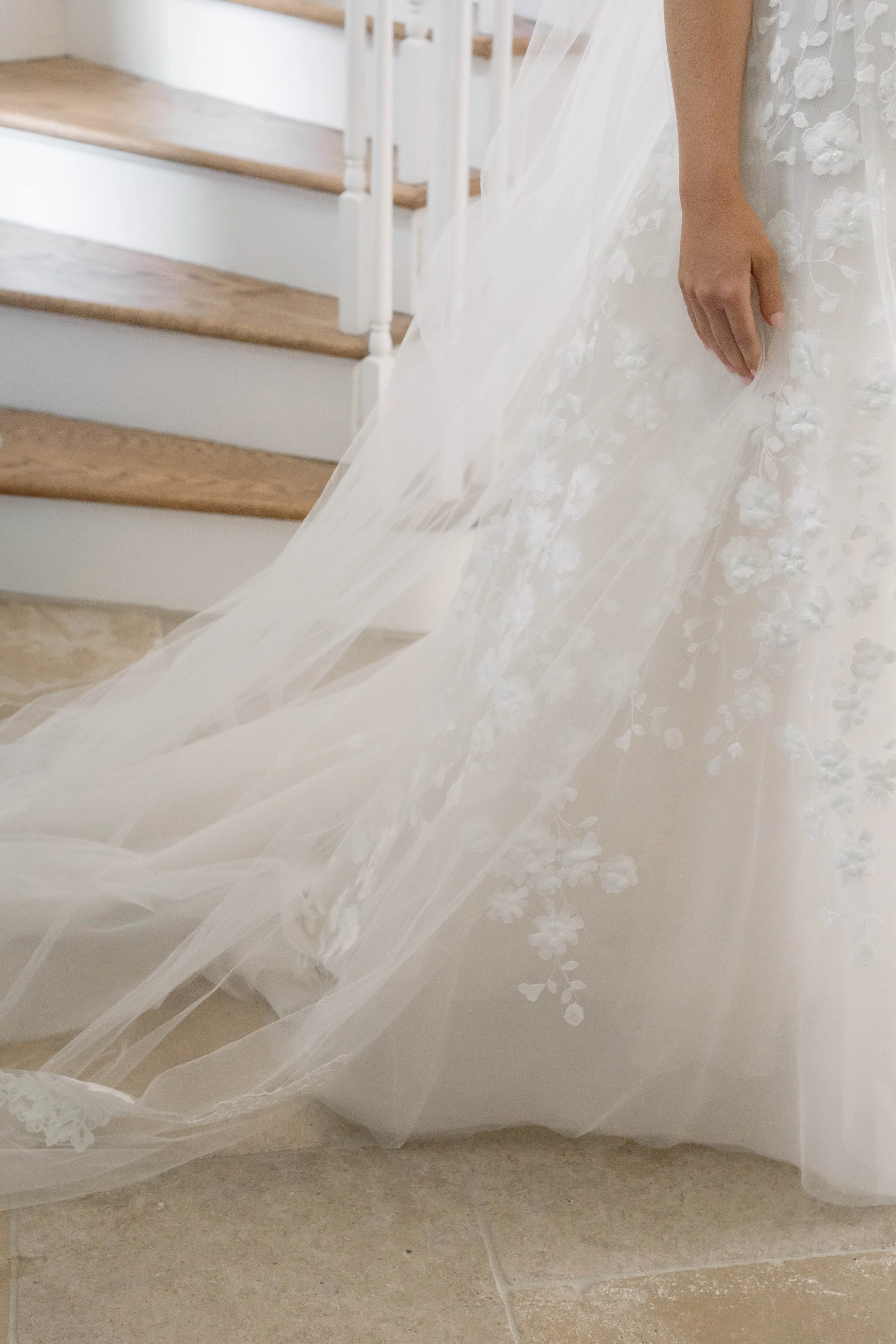 Close-up of a bride's hand resting on her wedding dress, showing detailed floral lace embroidery on the gown, with wooden stairs in the background.