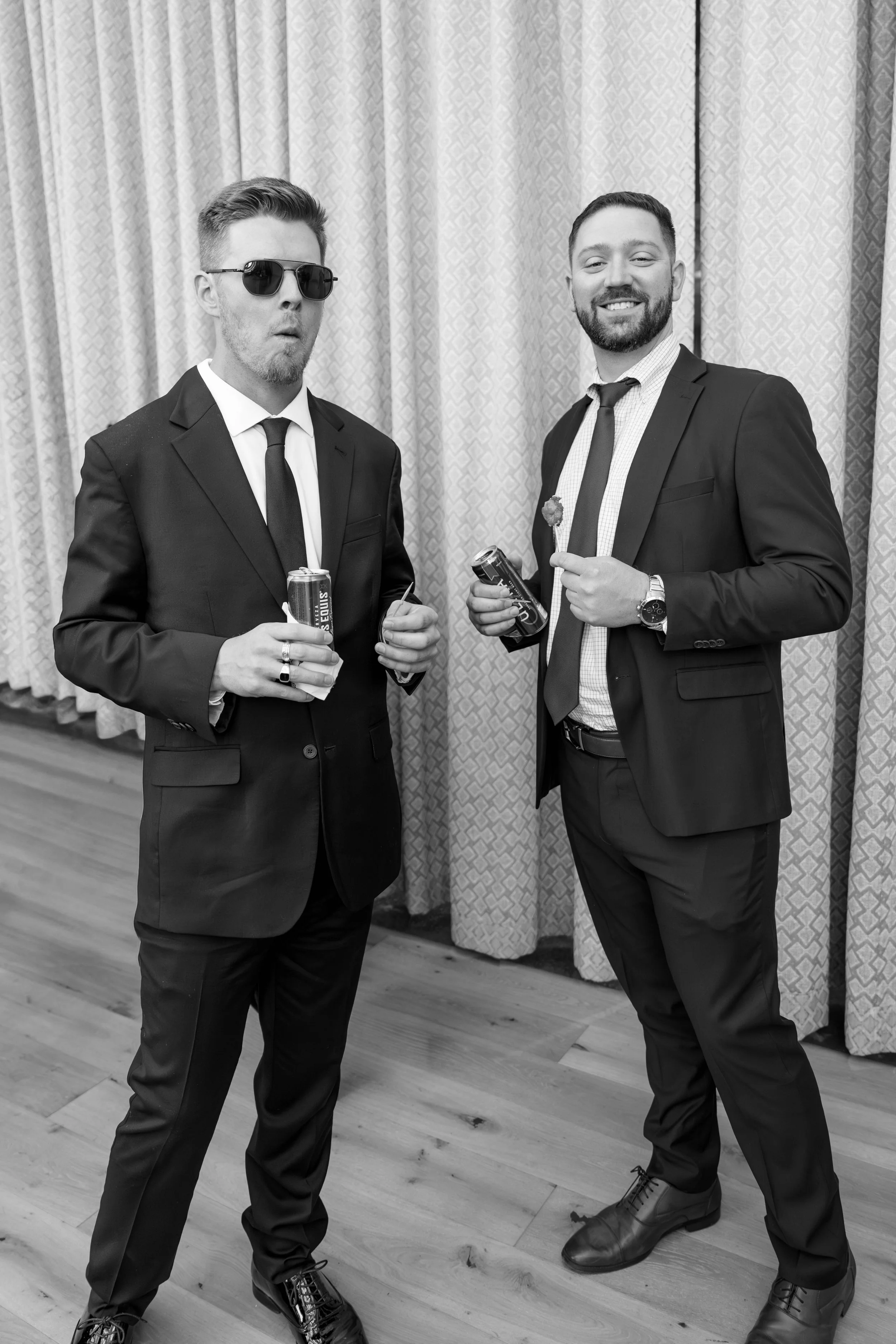 Two men in suits at an indoor event, one wearing sunglasses and holding a soda can, the other smiling and holding a soda can and a flower, standing in front of curtains.