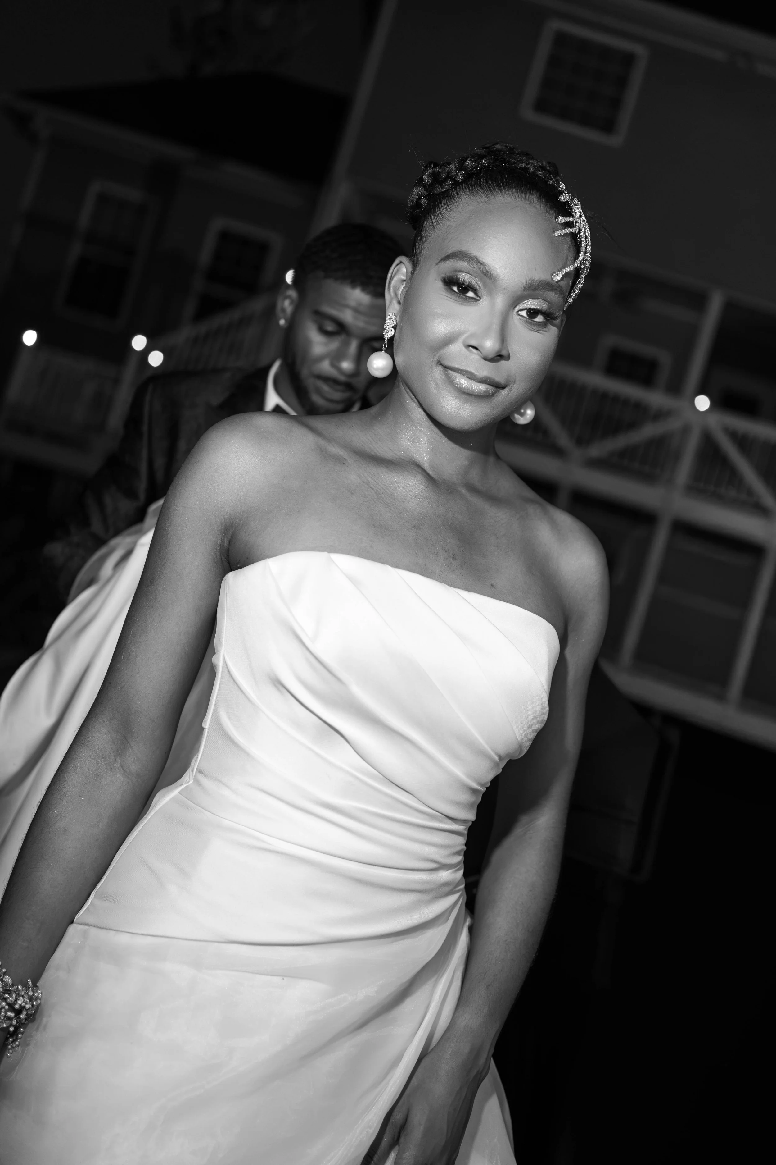 A woman dressed in a strapless, elegant gown with intricate detailing, wearing large pearl earrings and a beaded hair accessory. She has styled hair and a confident smile. In the background, a man in a suit is adjusting his tie, with a building and s