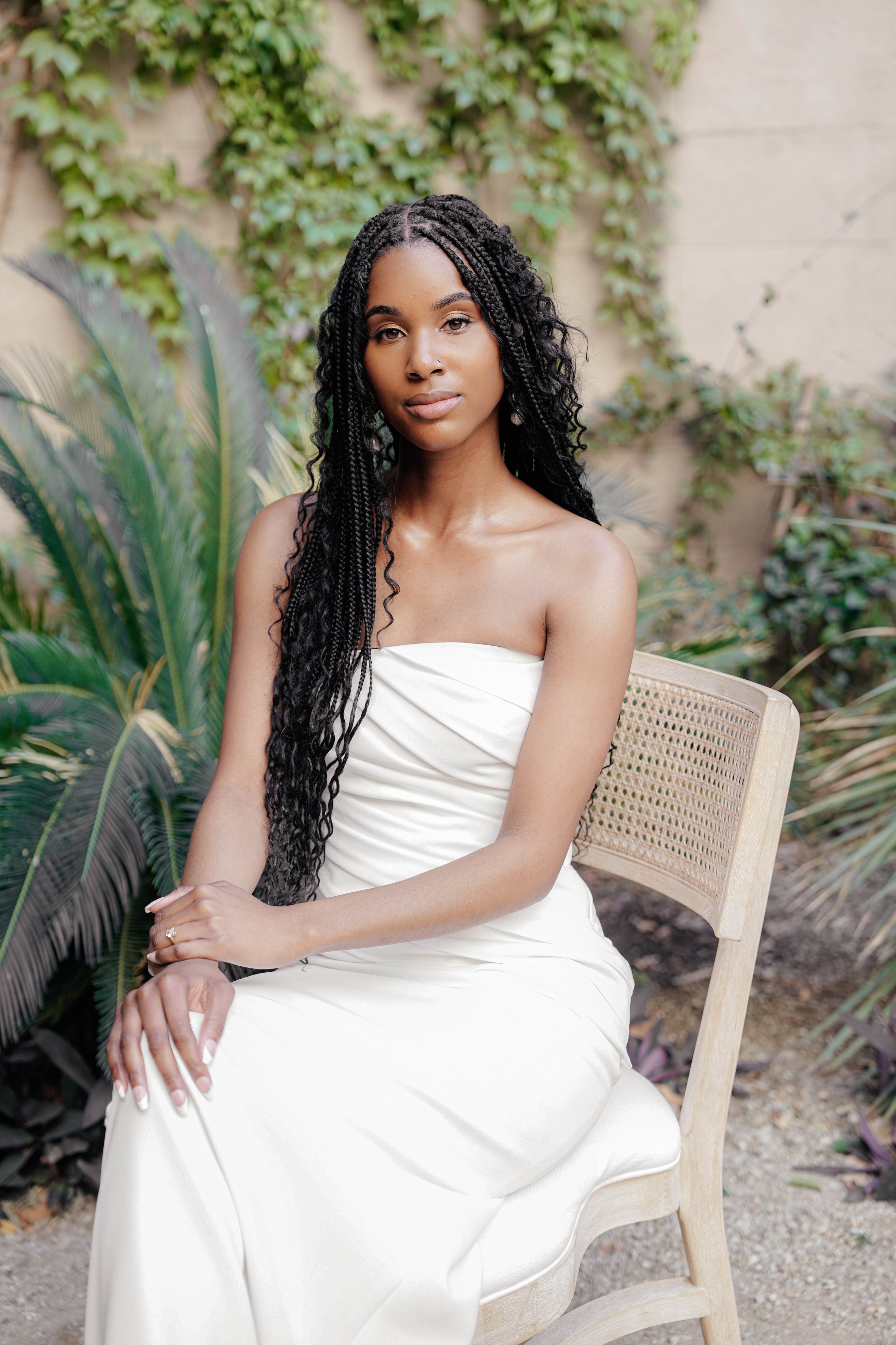 A young woman with long, braided black hair sitting on a wooden chair outdoors amidst green plants and ivy