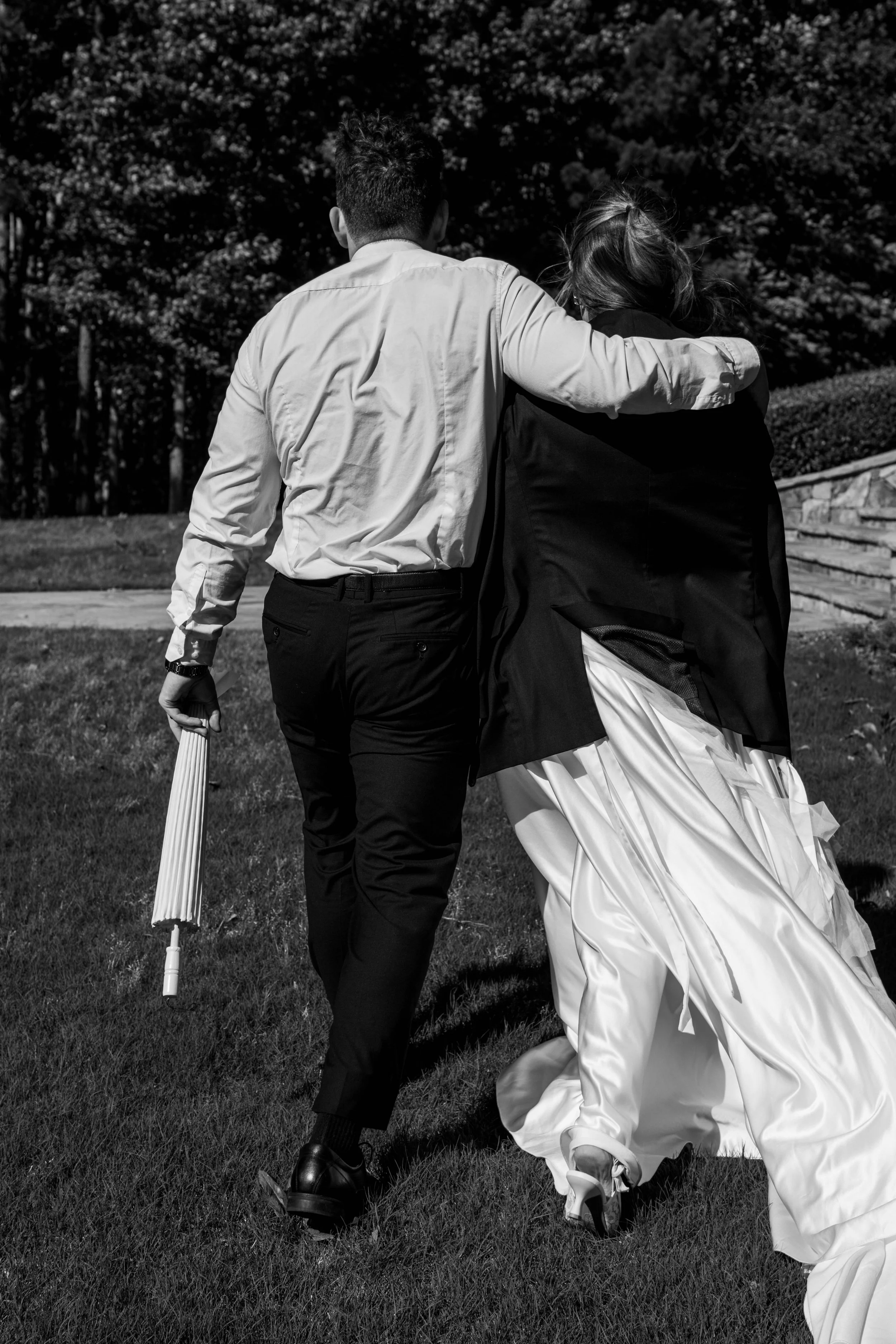 A man and a woman are walking together outdoors on grass, with the man having his arm around the woman. The man is dressed in a light-colored shirt and dark pants, holding a closed umbrella, and the woman is wearing a wedding dress. The scene is capt