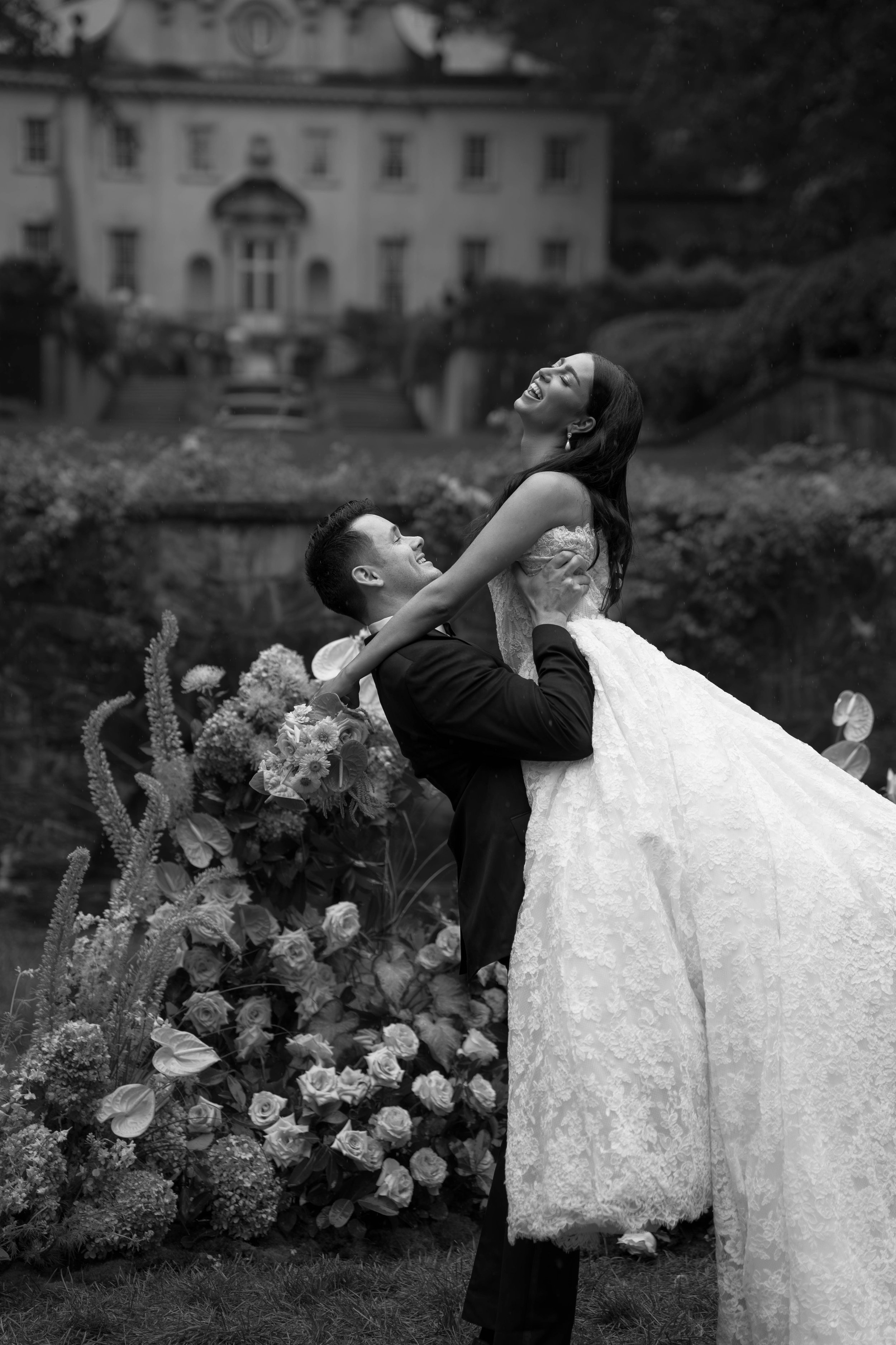 A black and white photo of a joyful bride and groom, with the groom lifting the bride, surrounded by flowers, in front of a large house with multiple windows and a staircase.