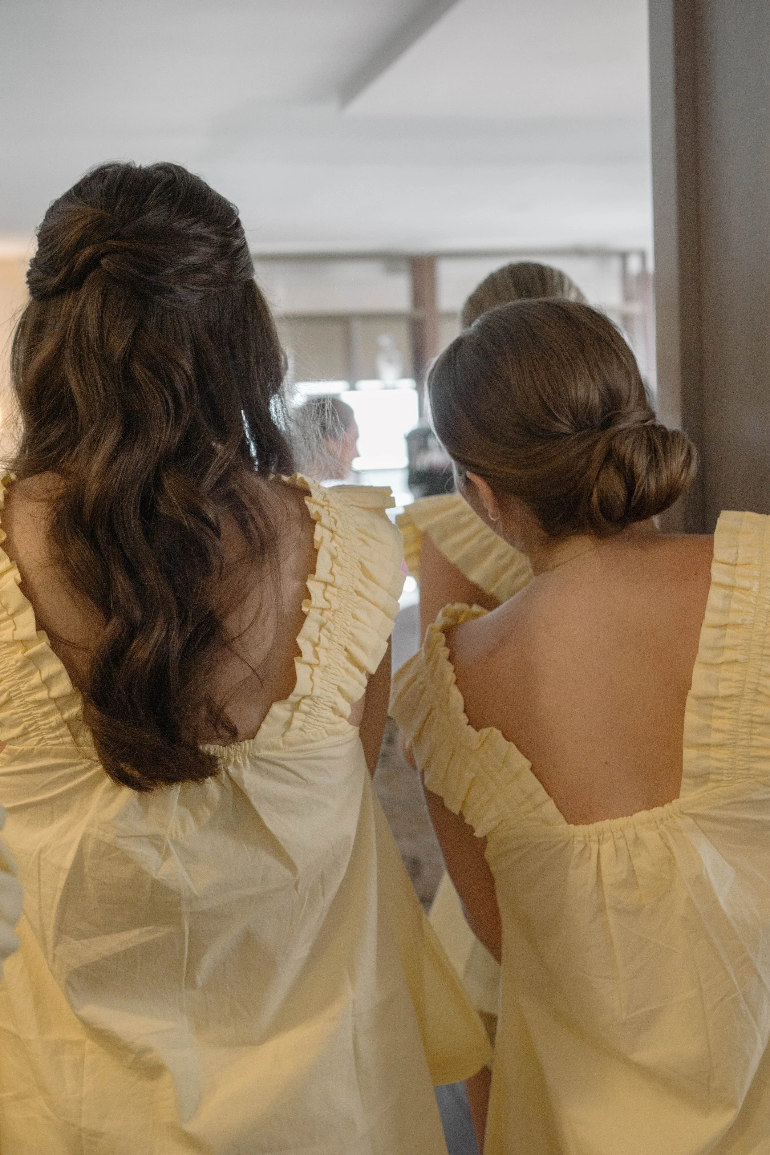 Back view of three women with styled hair, dressed in yellow gowns, likely preparing for a formal event or wedding.