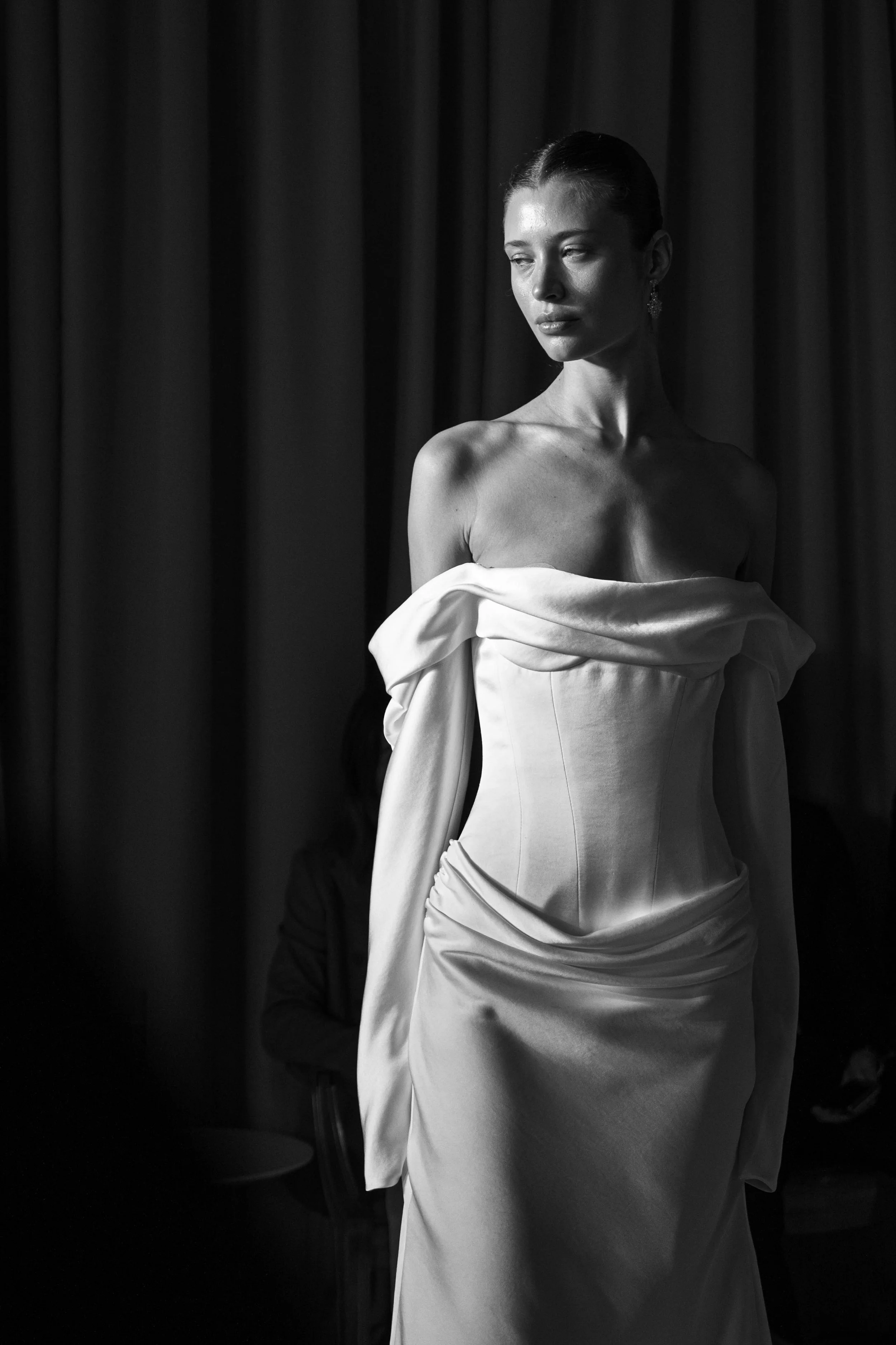 A black and white photo of a woman in a strapless dress standing in front of a dark curtain.