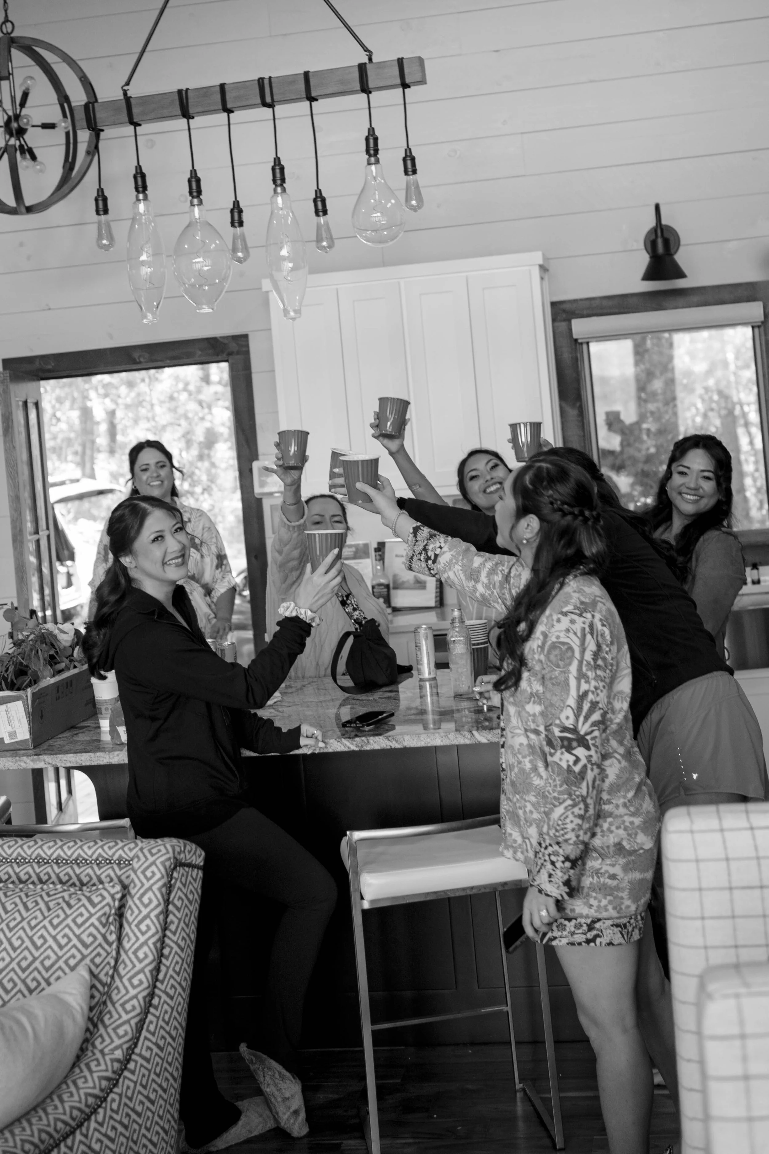 A group of women gathered in a kitchen, raising cups in a toast and smiling at each other. The kitchen has wooden walls, a window, and a modern light fixture with hanging bulbs.