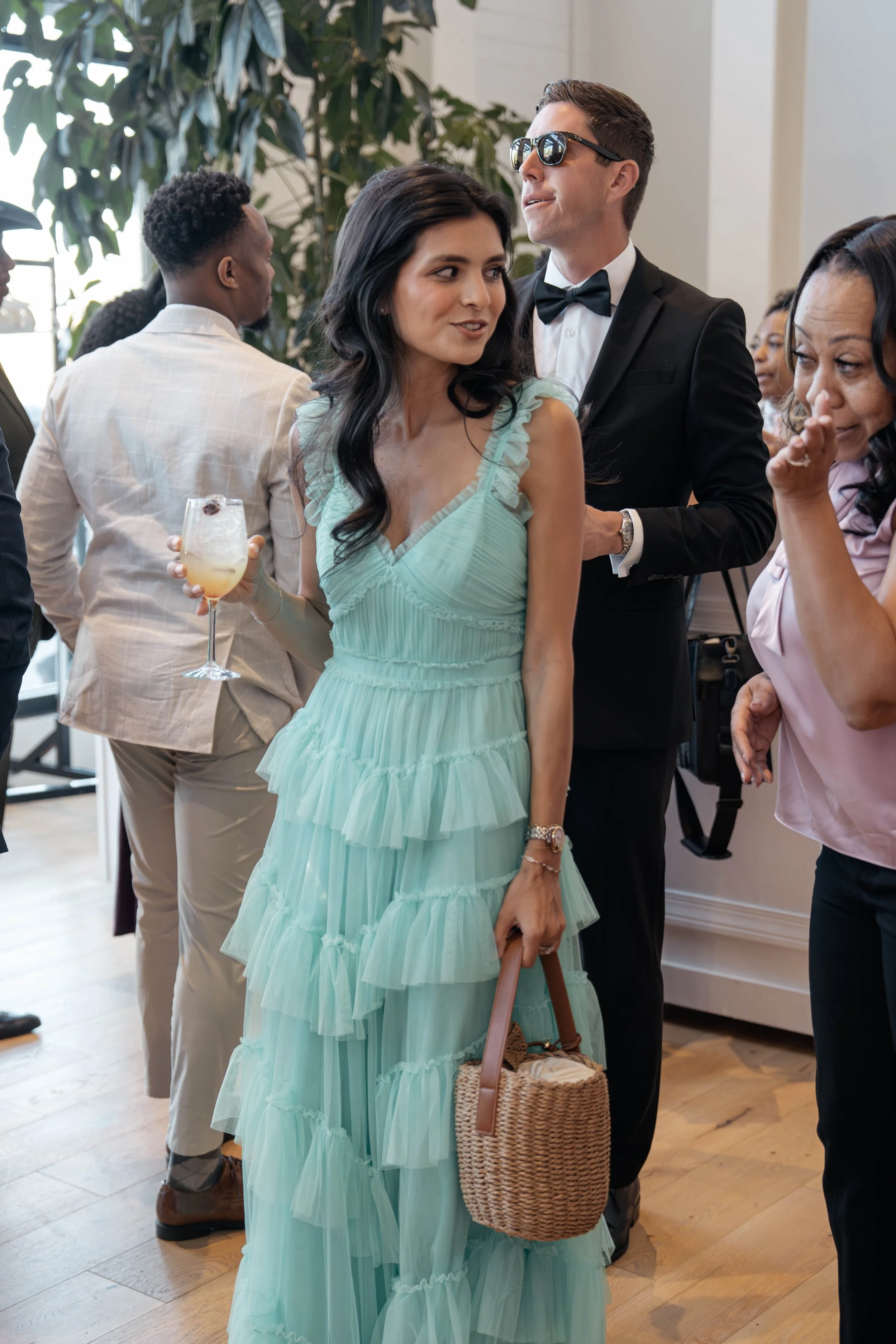 A woman in a light blue, ruffled dress holding a wicker basket and a drink, standing in a social gathering with people in formal attire.