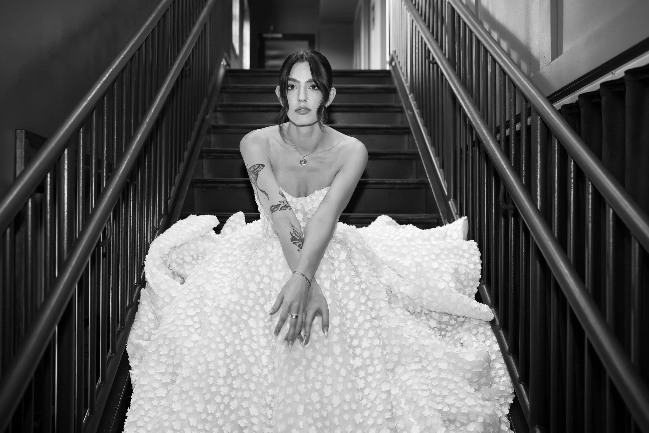 Black and white photo of a woman with dark hair sitting on a staircase in a strapless white dress.