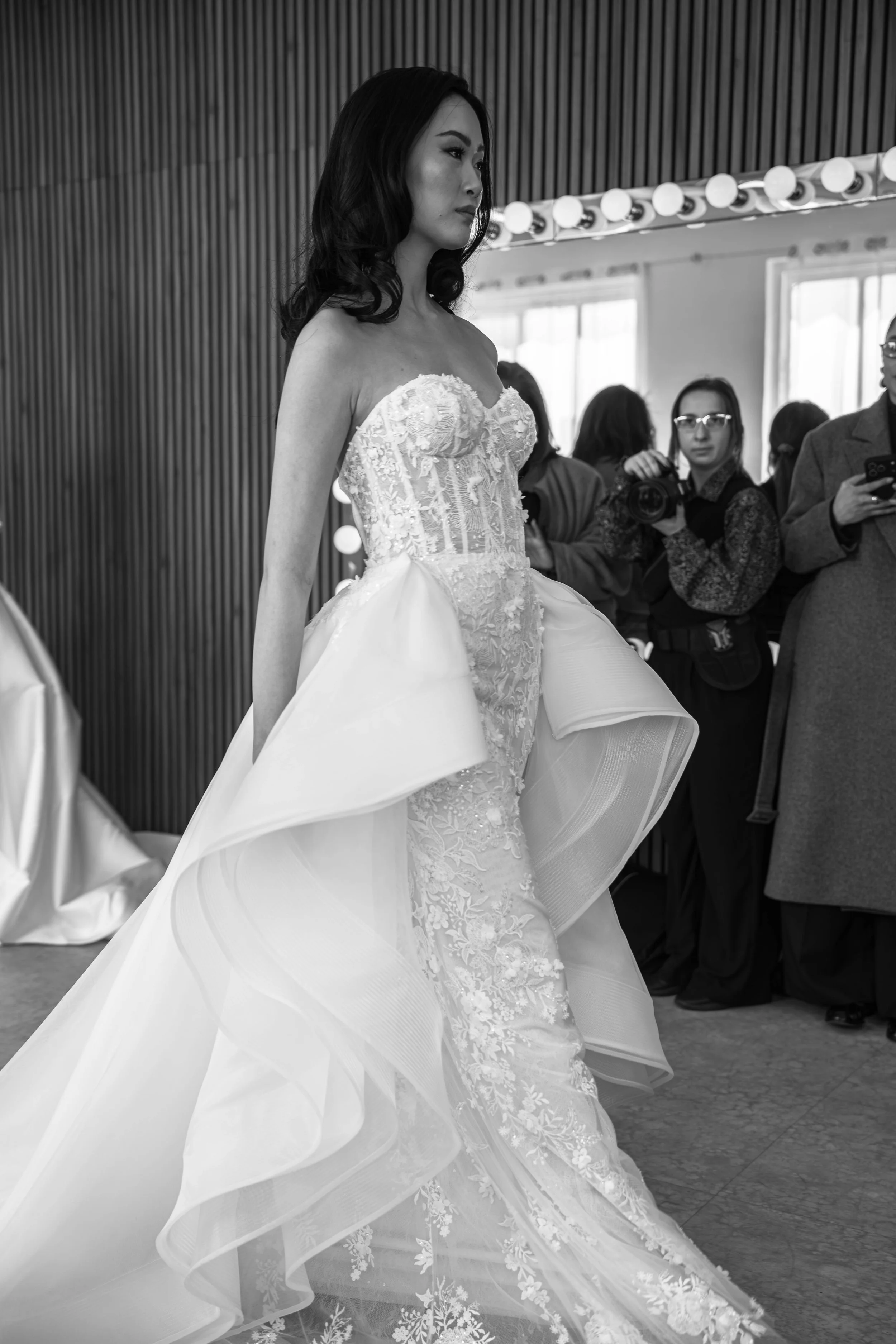 A woman in an ornate wedding dress with floral lace details stands indoors, surrounded by other people, some holding cameras or phones, in front of a wall with vertical wood slats and a row of light bulbs.