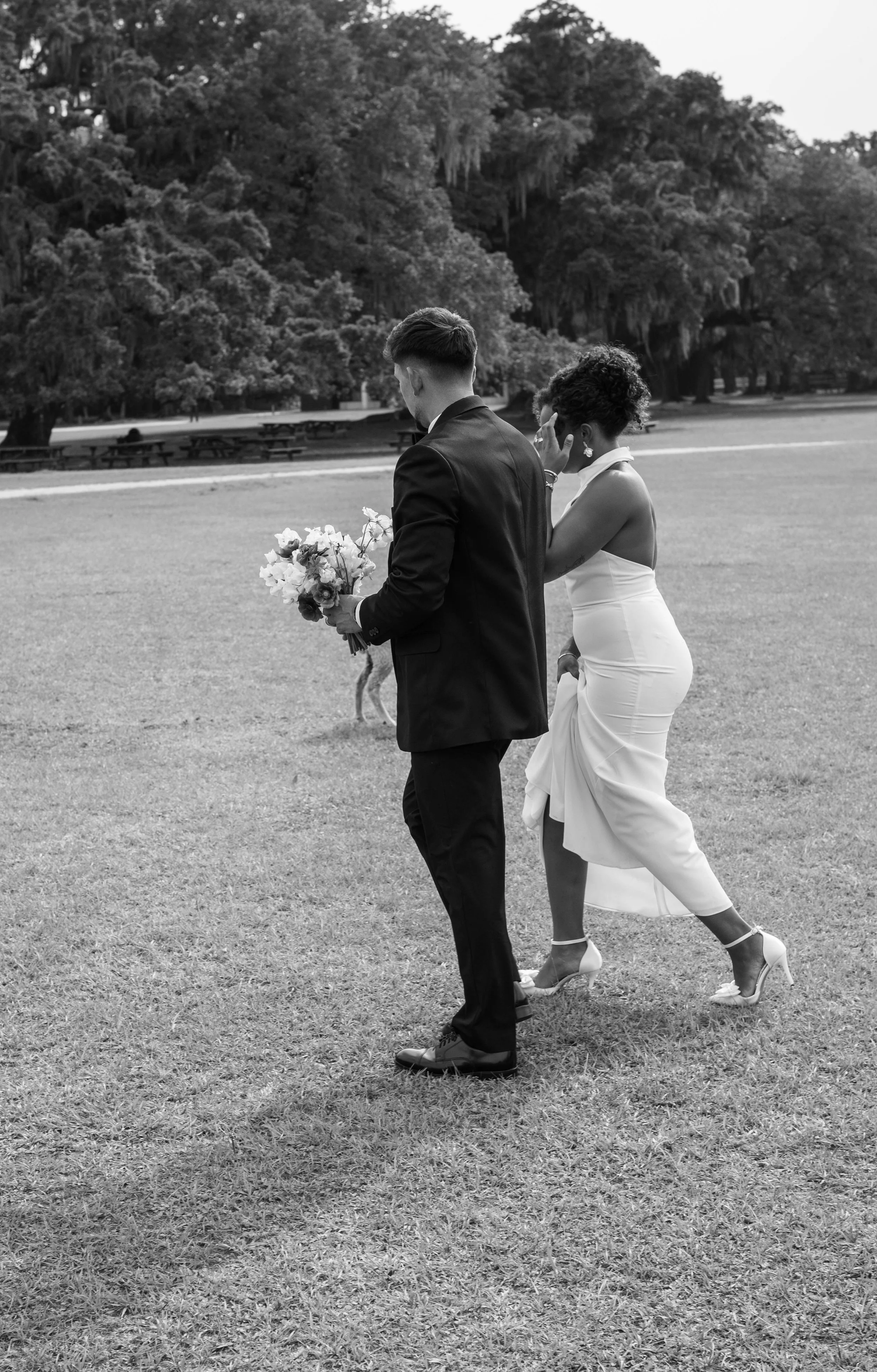 A black and white photo of a bride and groom walking in a park. The groom is holding a bouquet, and the bride appears emotional, walking behind him. Trees and picnic tables are visible in the background.