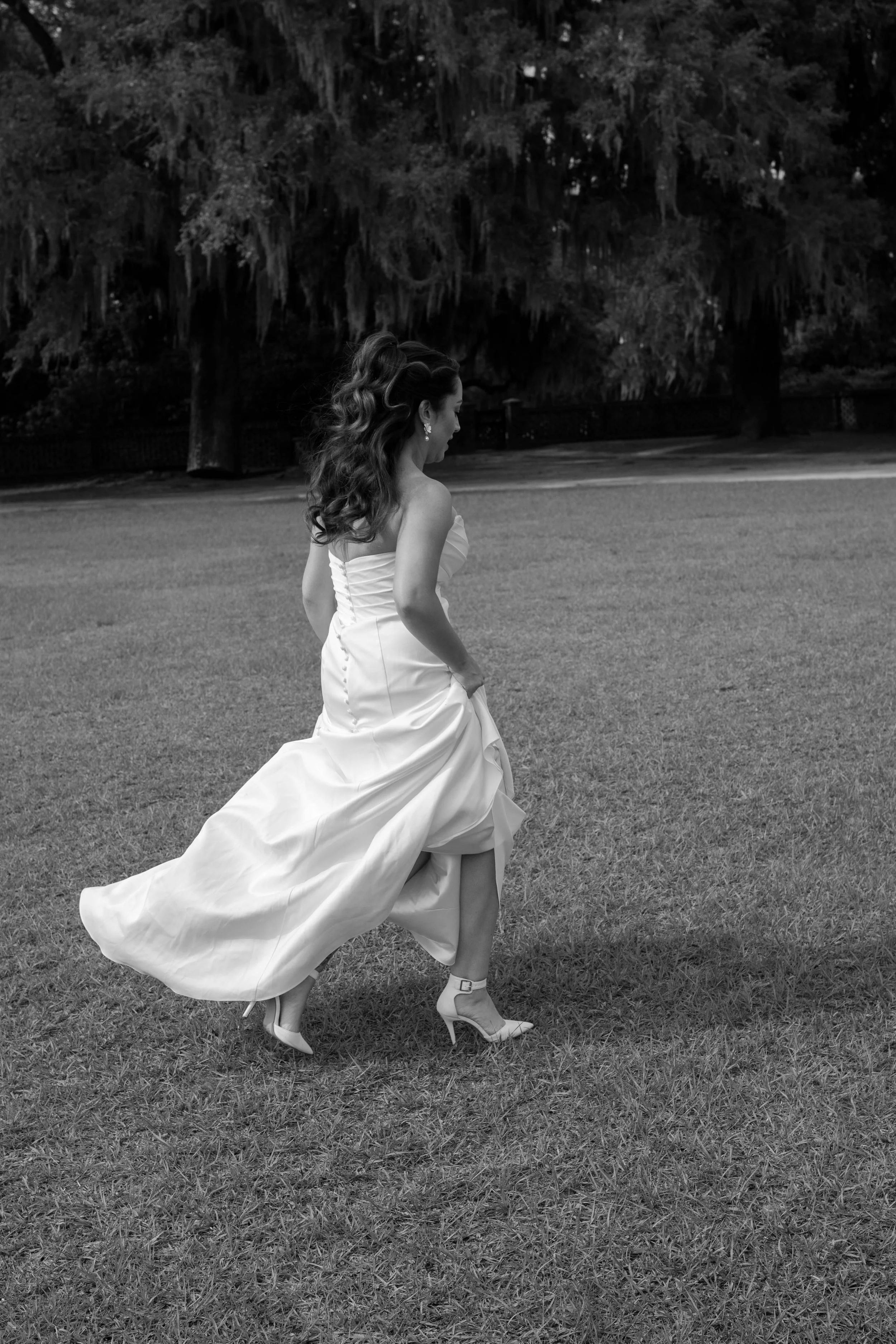 A woman in a wedding dress walking on grass with trees in the background, black and white photo.