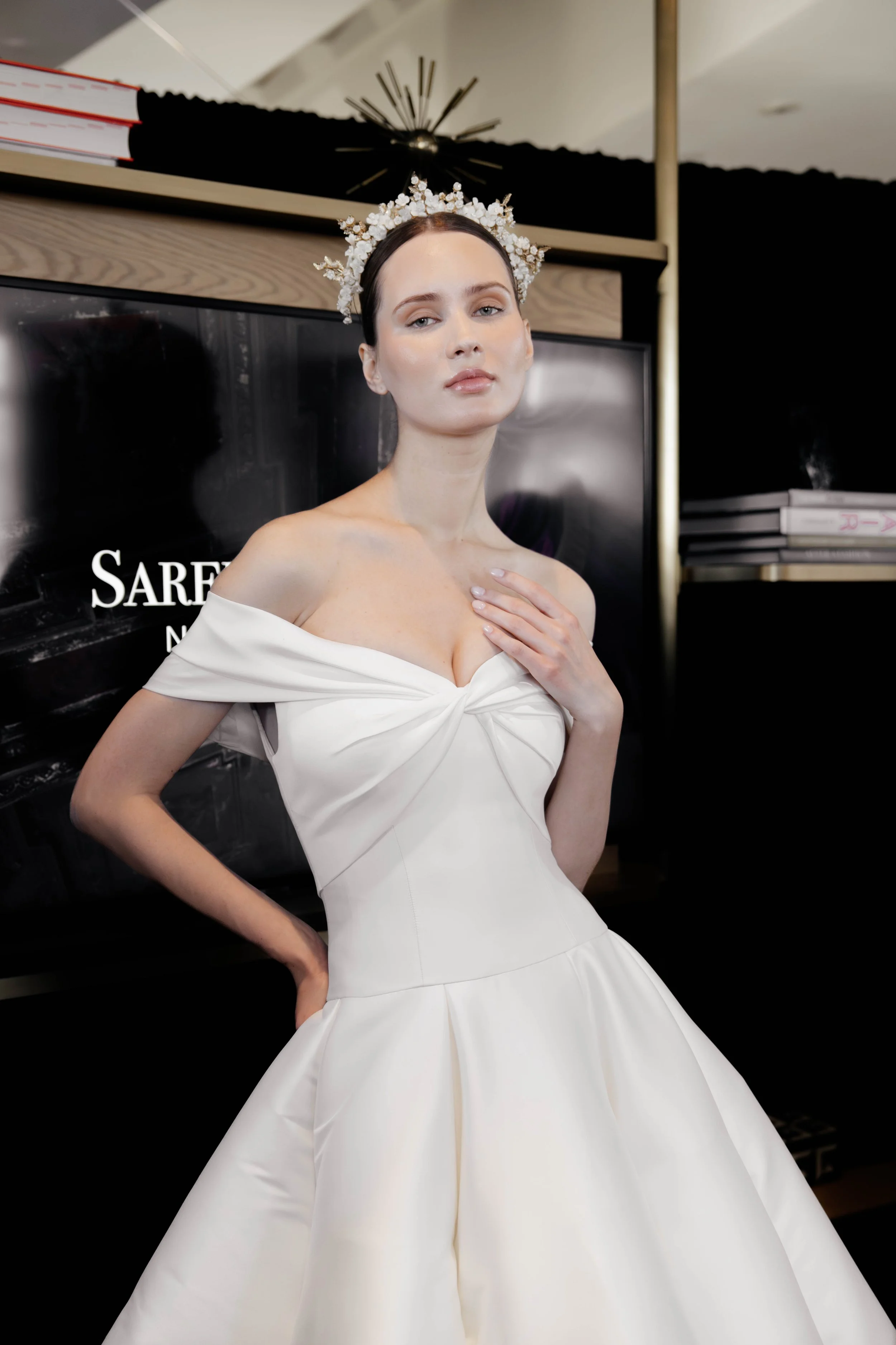 A woman in a white off-shoulder dress with a bow detail on the bodice, wearing a floral crown, posing indoors with a black background and a piano behind her.