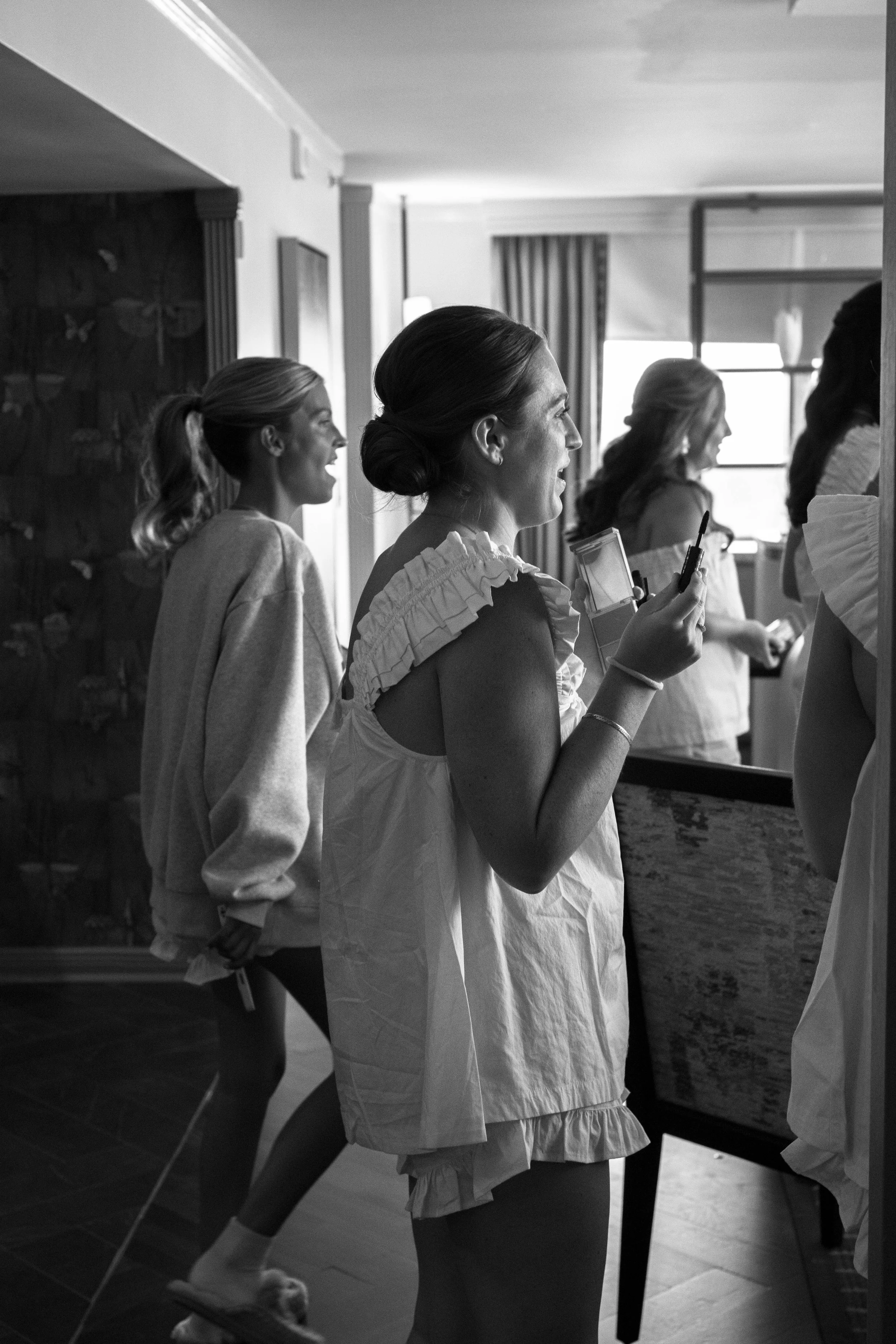 Women preparing in a room with makeup and mirrors, some applying makeup, others adjusting hair, with natural light coming through a window.