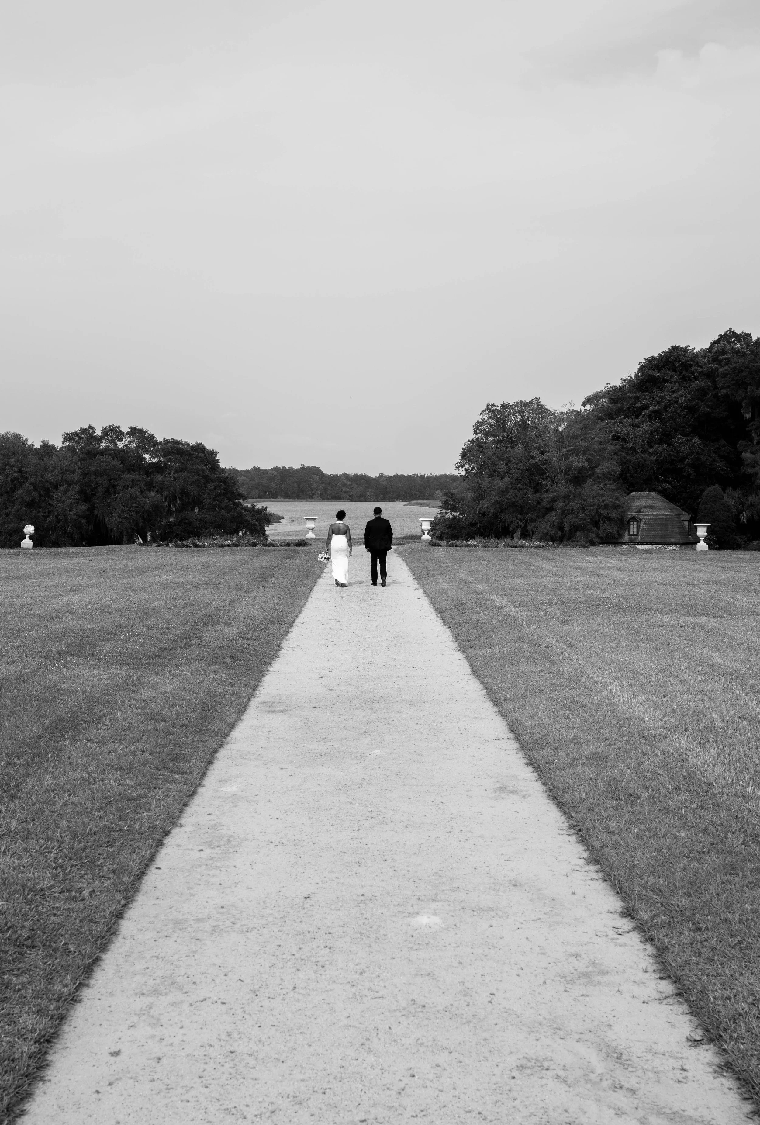 A black and white photo of a couple walking down a path in a large open field, surrounded by trees, with a distant body of water and a sky with minimal clouds in the background.