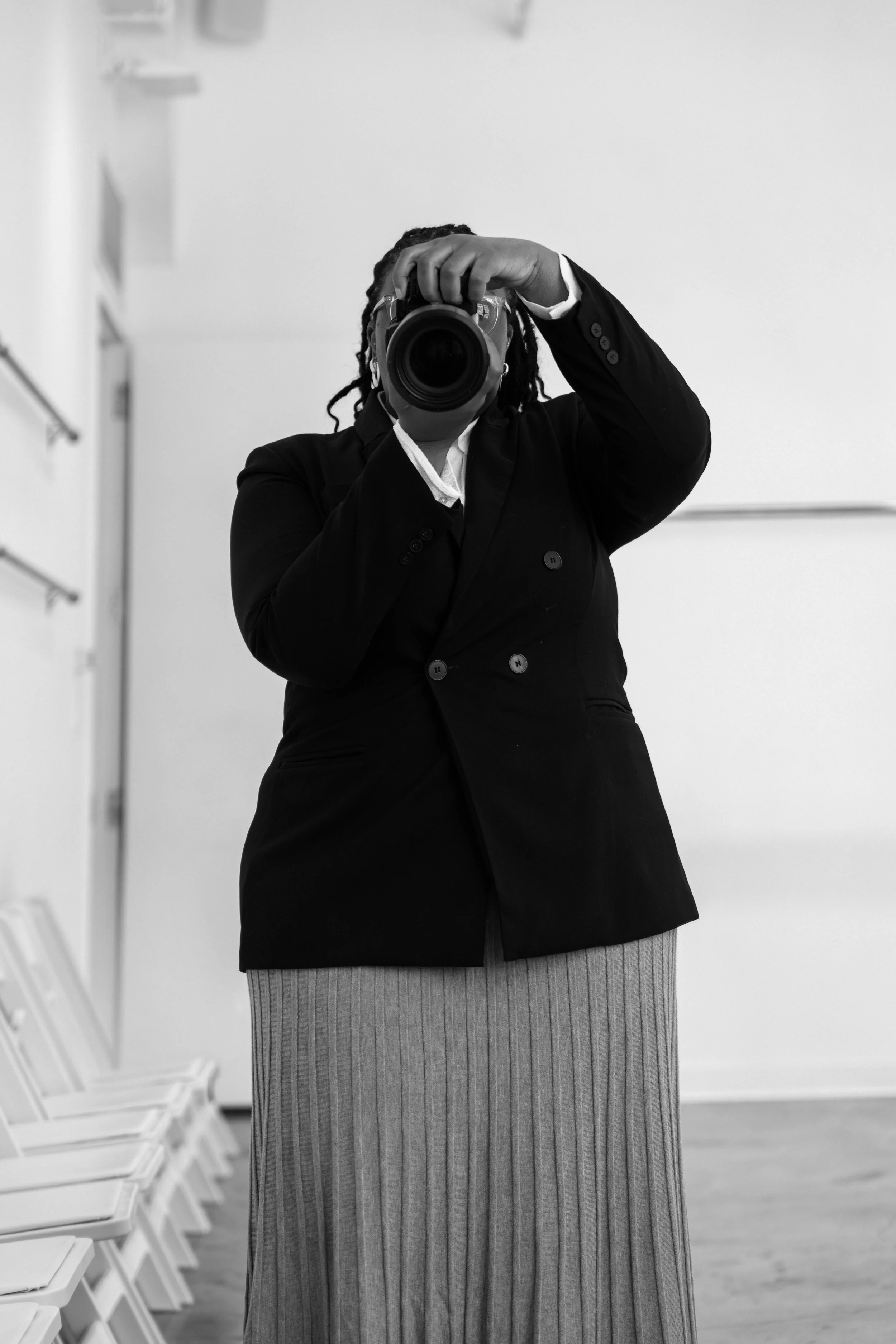 Person taking self-portrait with camera in a room with white walls, white chairs stacked on the side, wearing a dark blazer and striped skirt. Black and white photo.