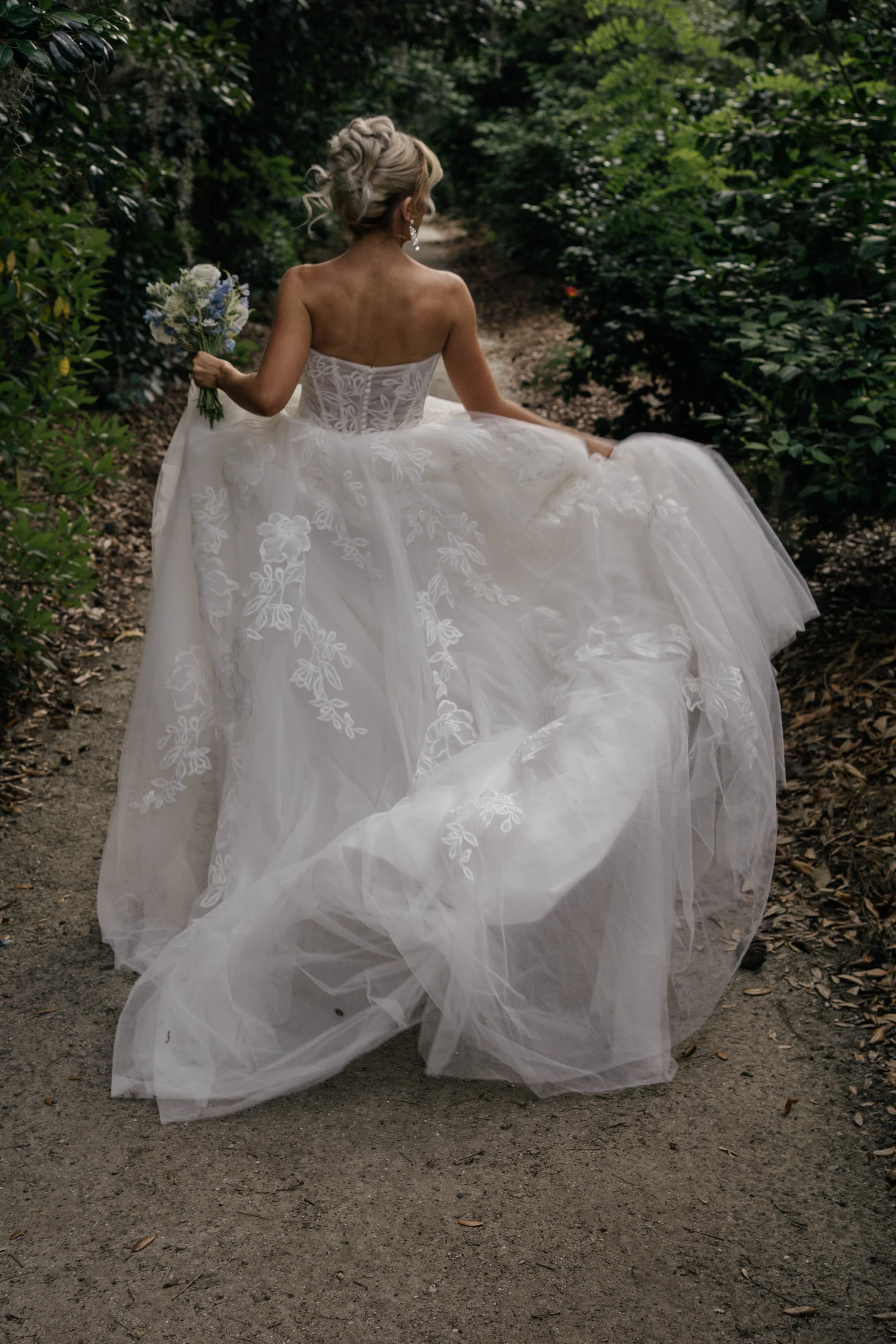 A bride in a strapless, embroidered wedding gown walking on a wooded path while holding a bouquet of flowers.