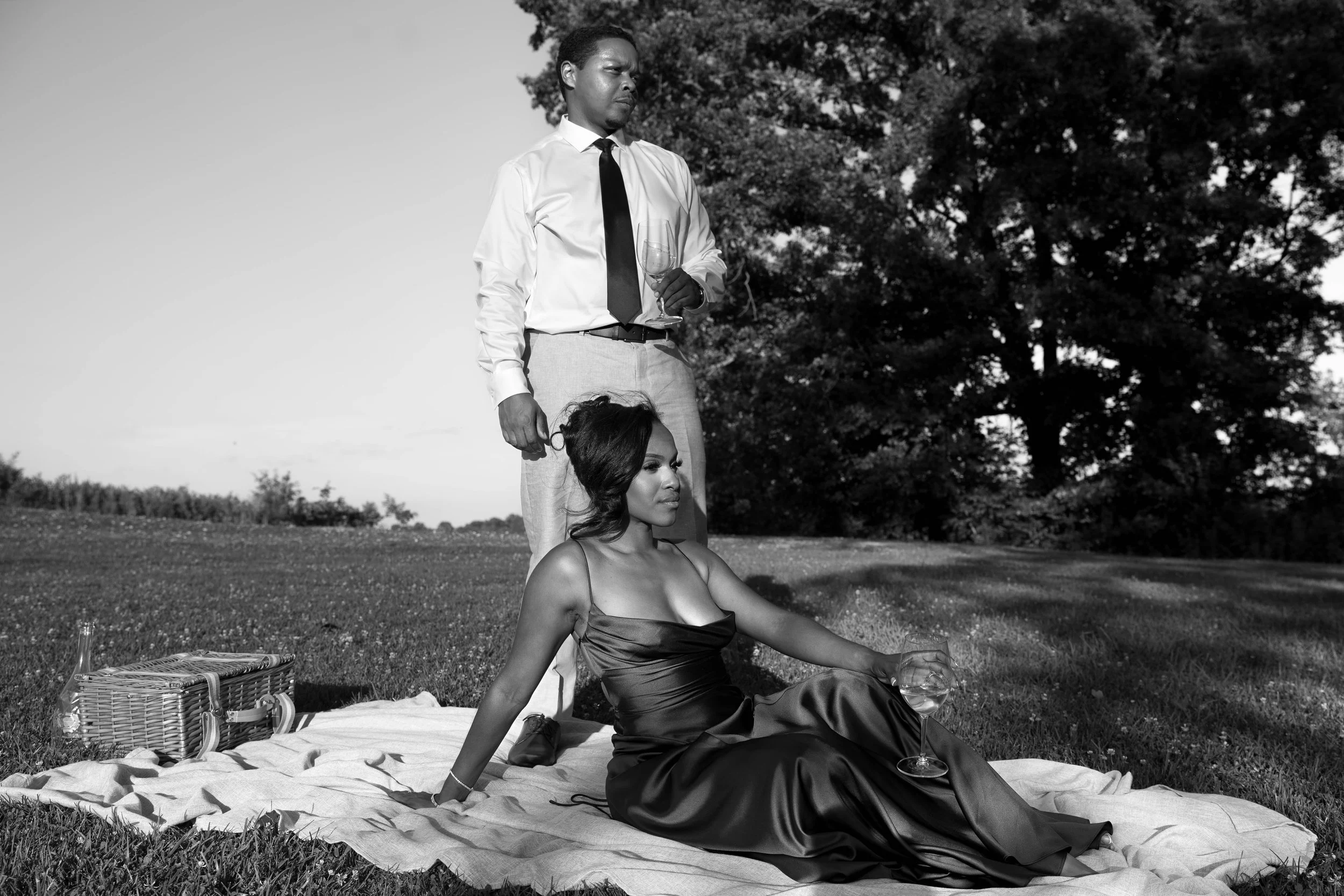 A black and white photo of a man standing behind a seated woman on a blanket in a park, holding a wine glass. The woman is wearing a satin dress and sitting with her arm extended, holding a wine glass, while the man is dressed in a shirt and tie, loo