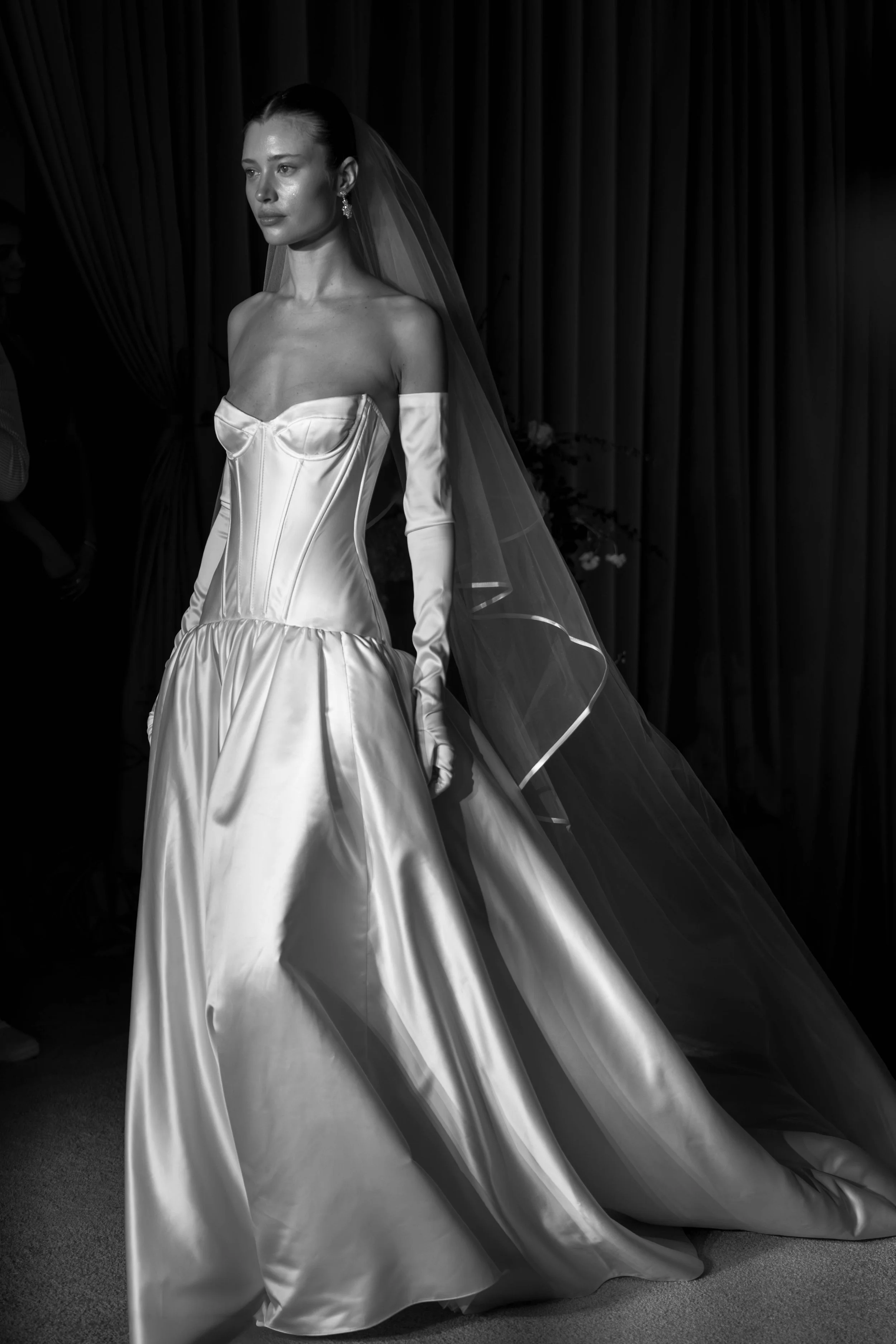 A woman in a wedding dress with long sleeves and a full skirt, wearing a veil, standing indoors in front of dark curtains.