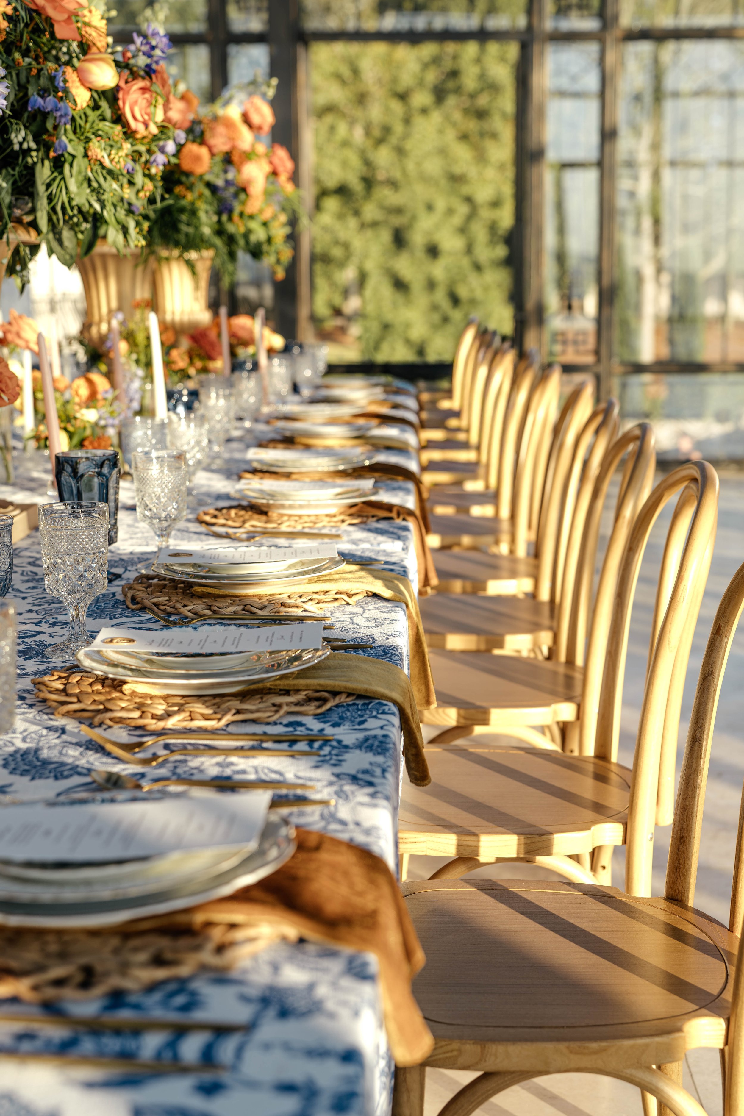 A long dining table set for a meal with elegant glassware, plates, and cutlery, decorated with floral arrangements in a sunlit greenhouse with large windows and wooden chairs along the table.