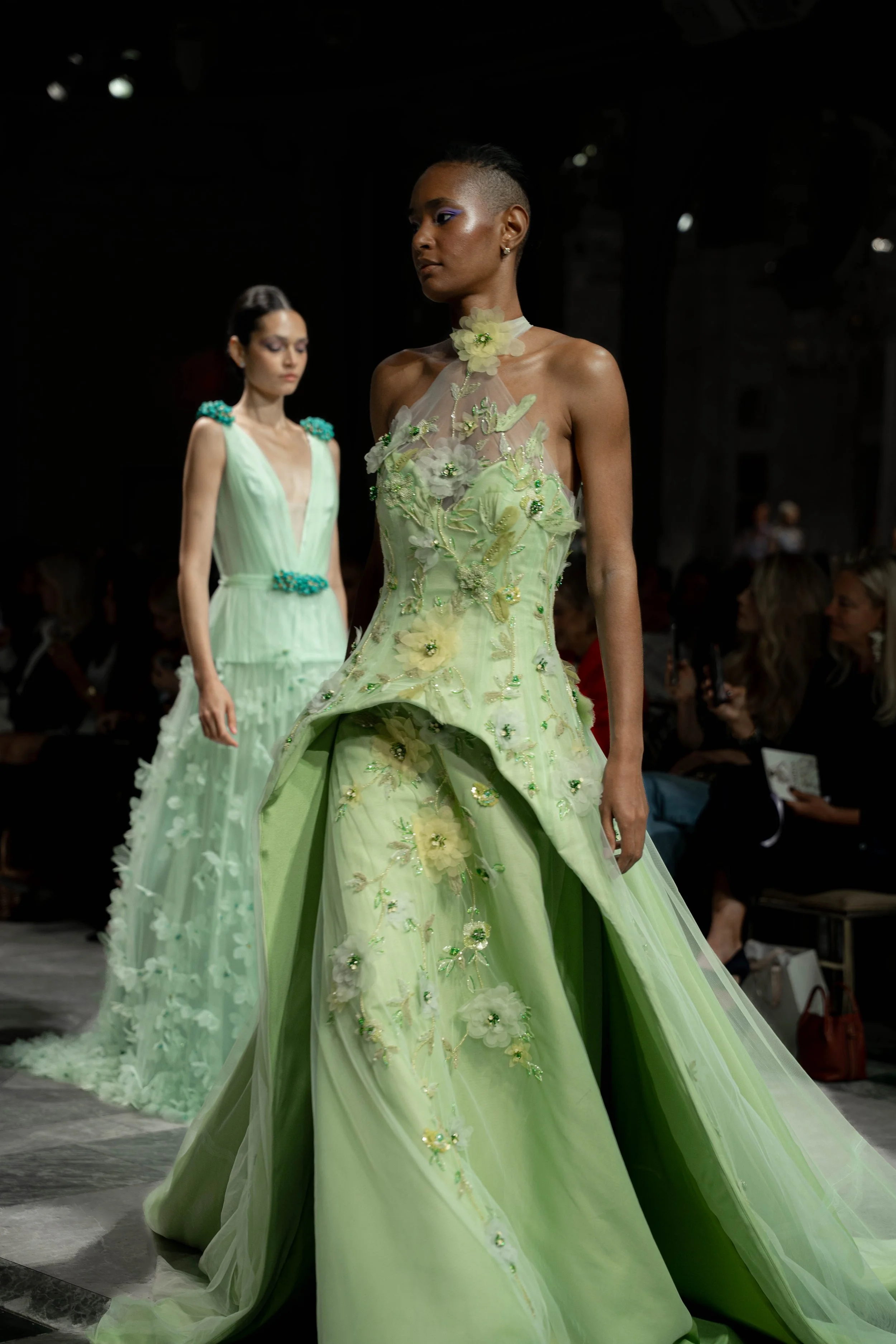 Two models on a runway, wearing elaborate green gowns with floral embroidery and 3D floral appliqués, during a fashion show.