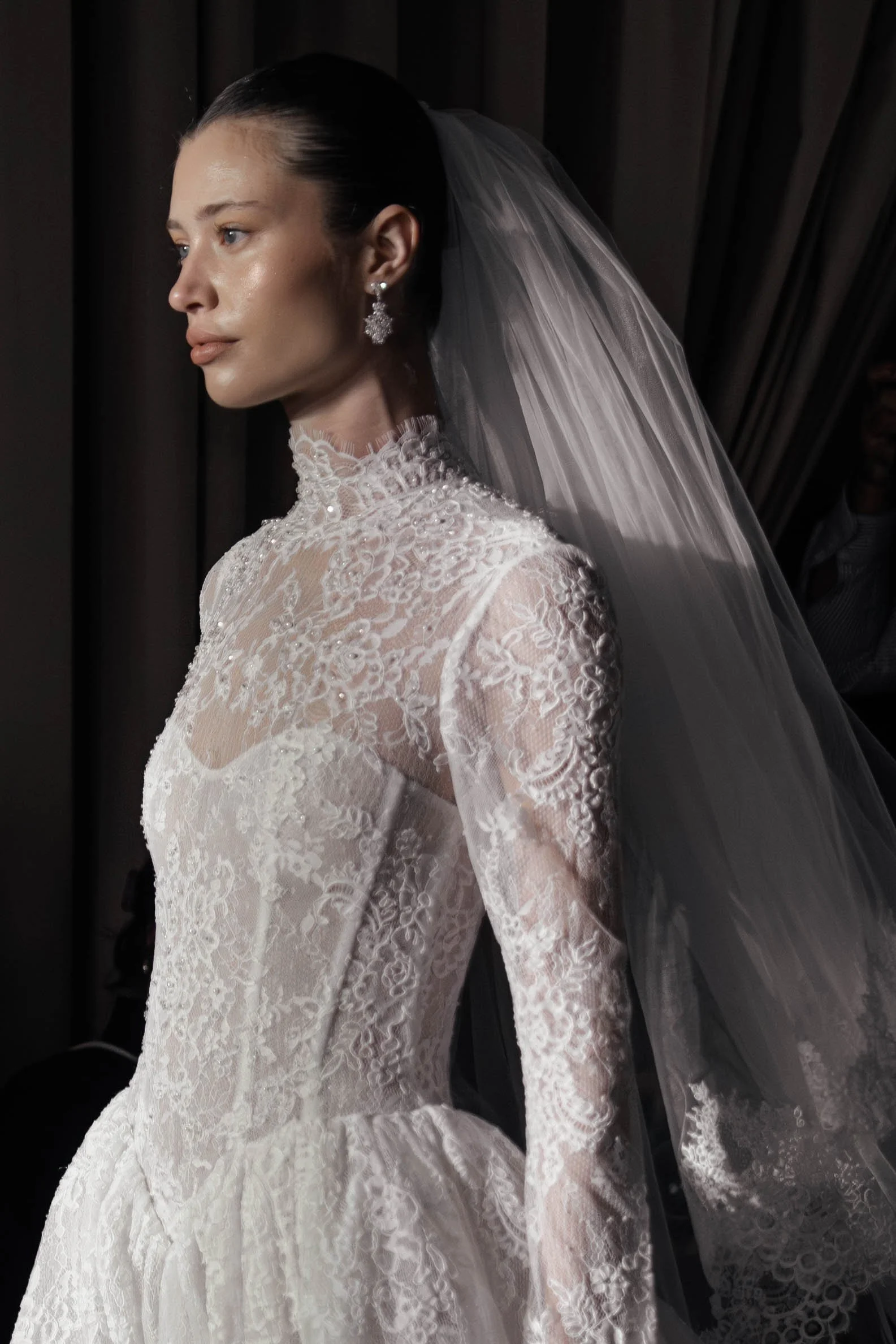 A woman in a white lace wedding dress with a high neckline, long sleeves, and floral lace embroidery, wearing a veil and sparkling chandelier earrings, posing against a dark background.
