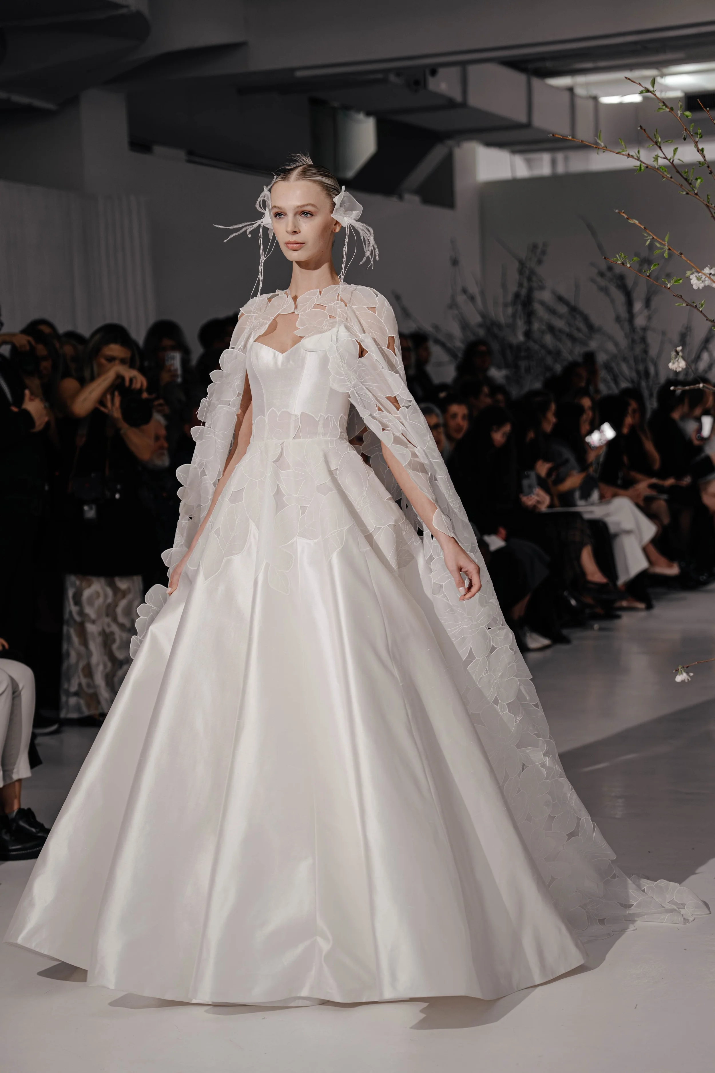 A model wearing a white wedding gown with floral details on the fabric walking on the runway at a fashion show. The audience is seated along the runway, some taking photos.