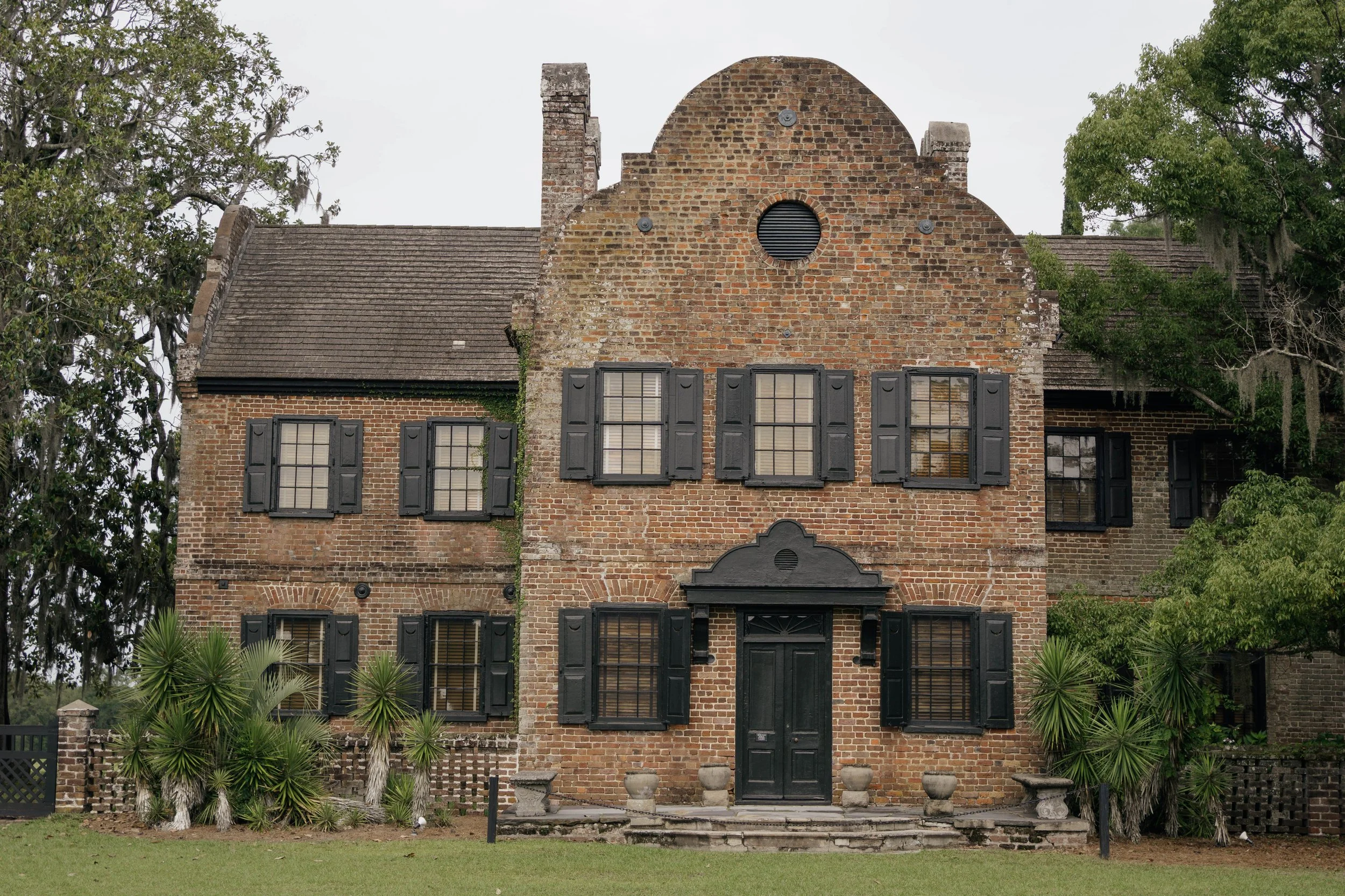 Historical brick house with black shutters, surrounded by greenery, and a front porch with steps and potted plants.