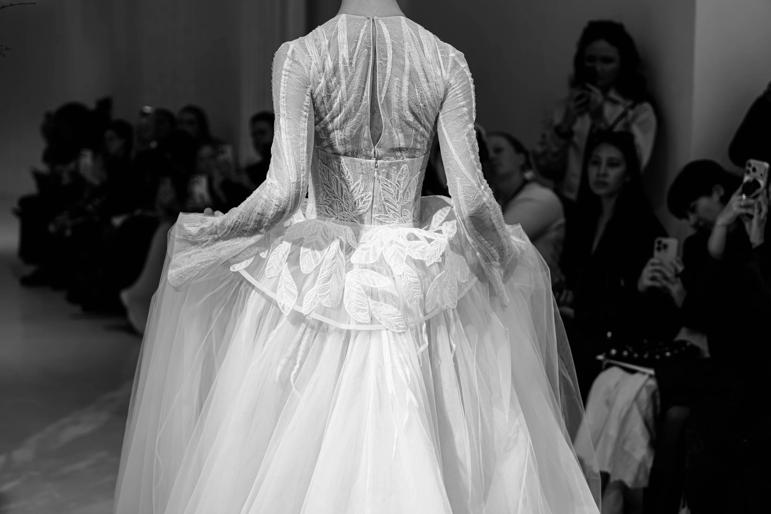 A person wearing an intricate, lace wedding dress with floral embroidery, walking on a runway during a fashion show, with seated audience members watching and taking photos.