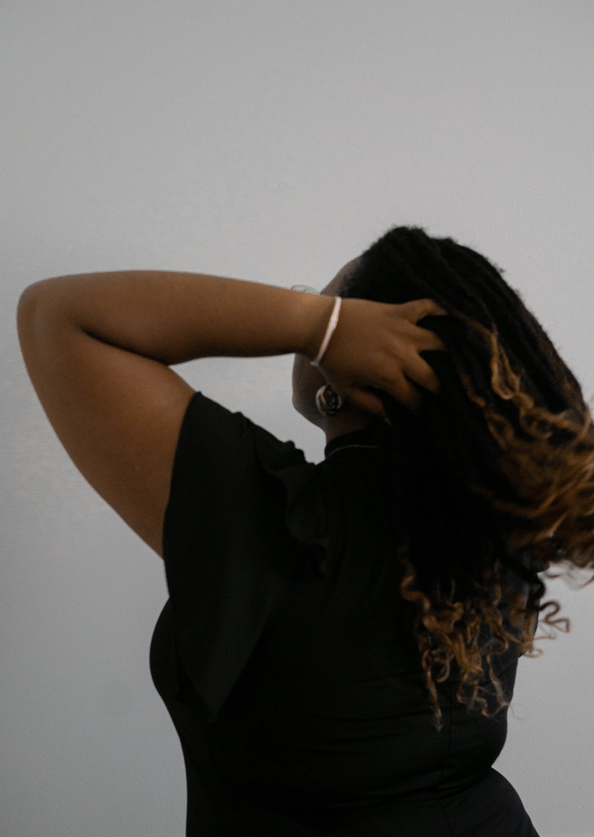 A woman with long curly hair, wearing a black shirt and earrings, holding her head with one hand, with a plain gray background.