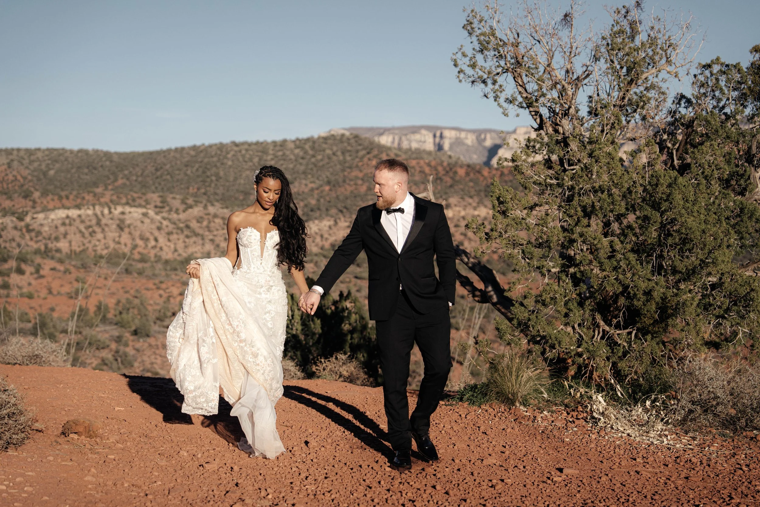 A bride and groom holding hands and walking outdoors in a desert landscape with mountains, trees, and clear blue sky.