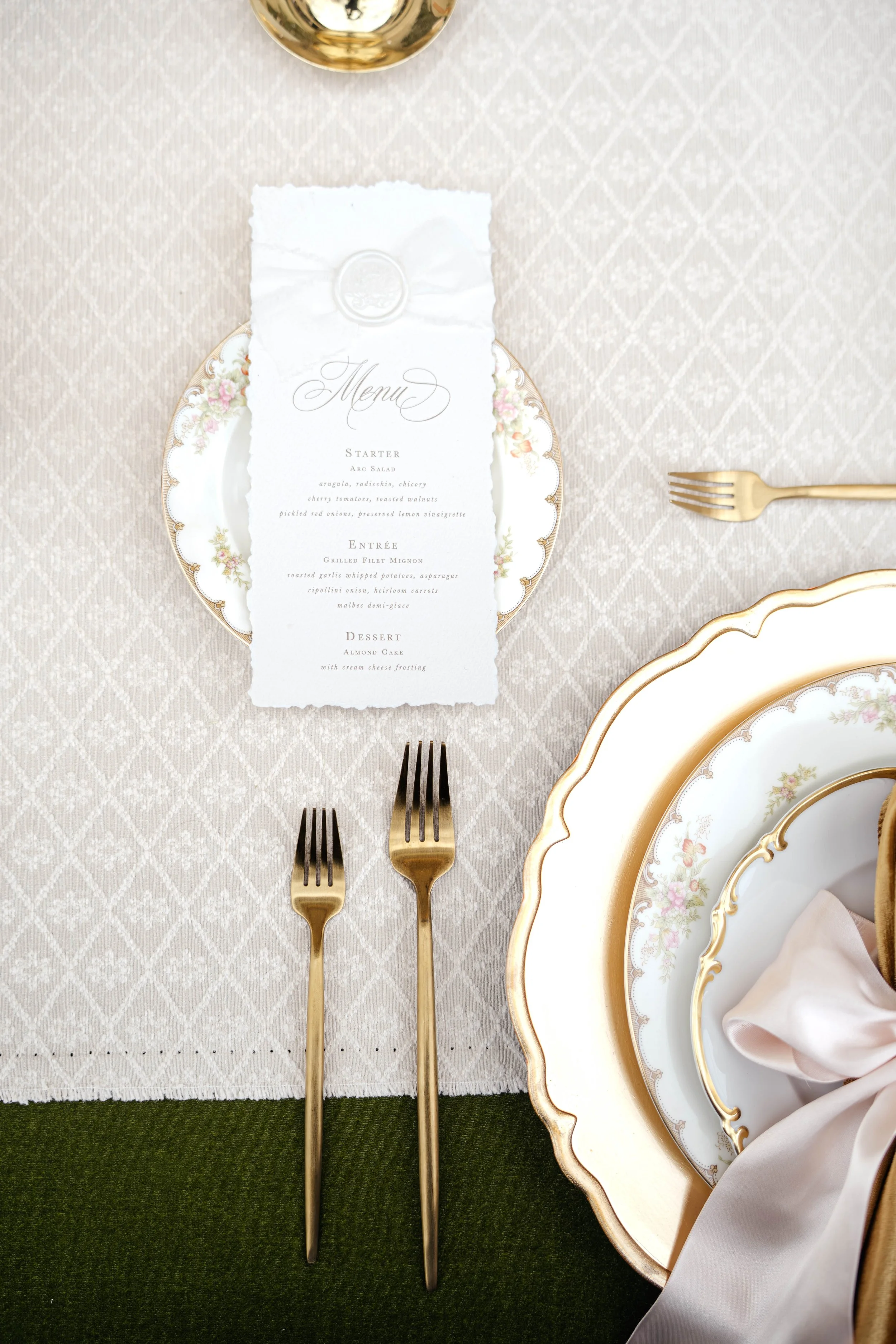 Elegant table setting with three gold forks, a floral-patterned plate, a menu on a torn white paper, a gold-rimmed plate, and a glass of white wine on a white textured tablecloth, with a dark green placemat under the utensils.