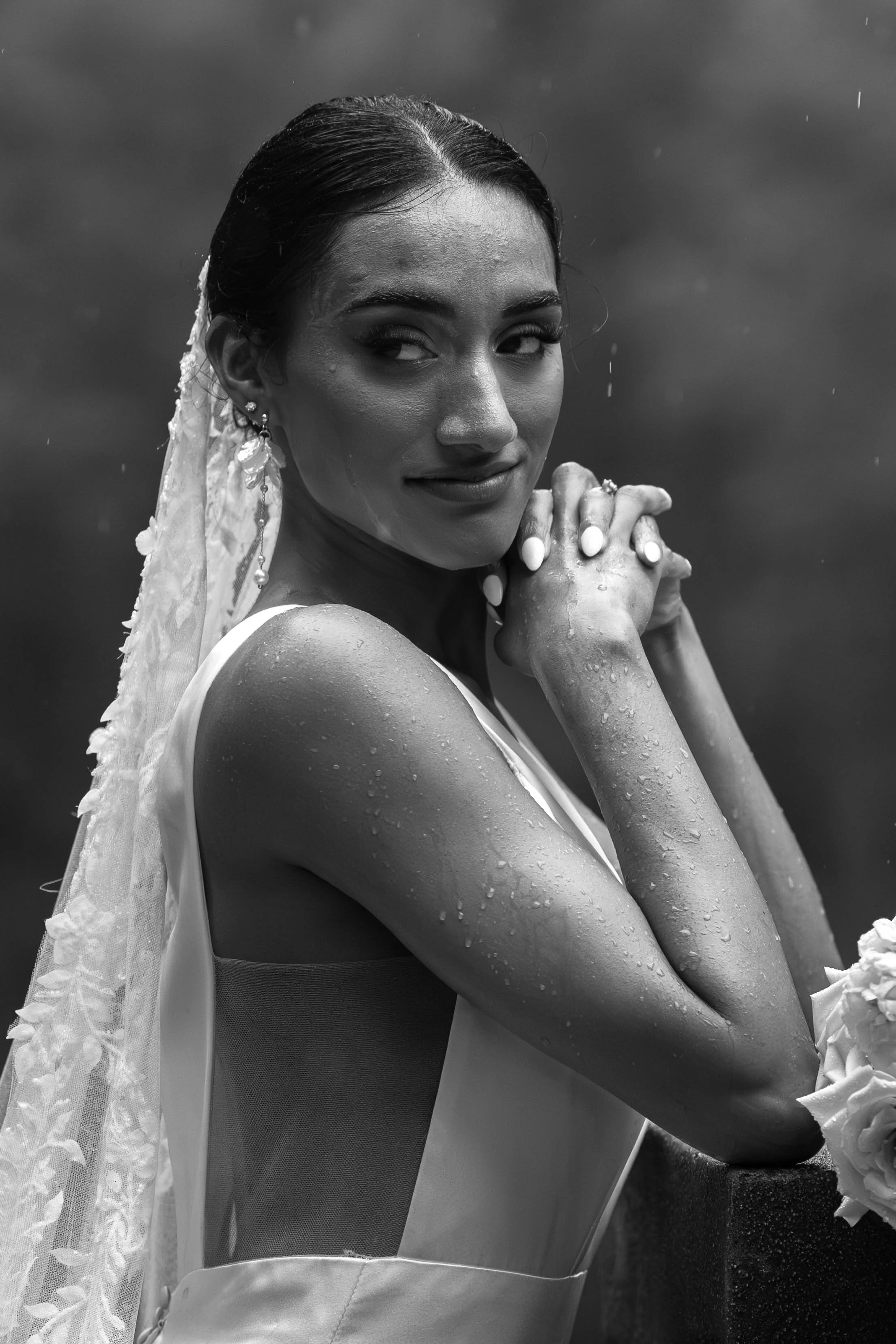 A woman with wet hair and water droplets on her skin, posing with her hands behind her head, wearing earrings and a dress with lace details, against a dark background.